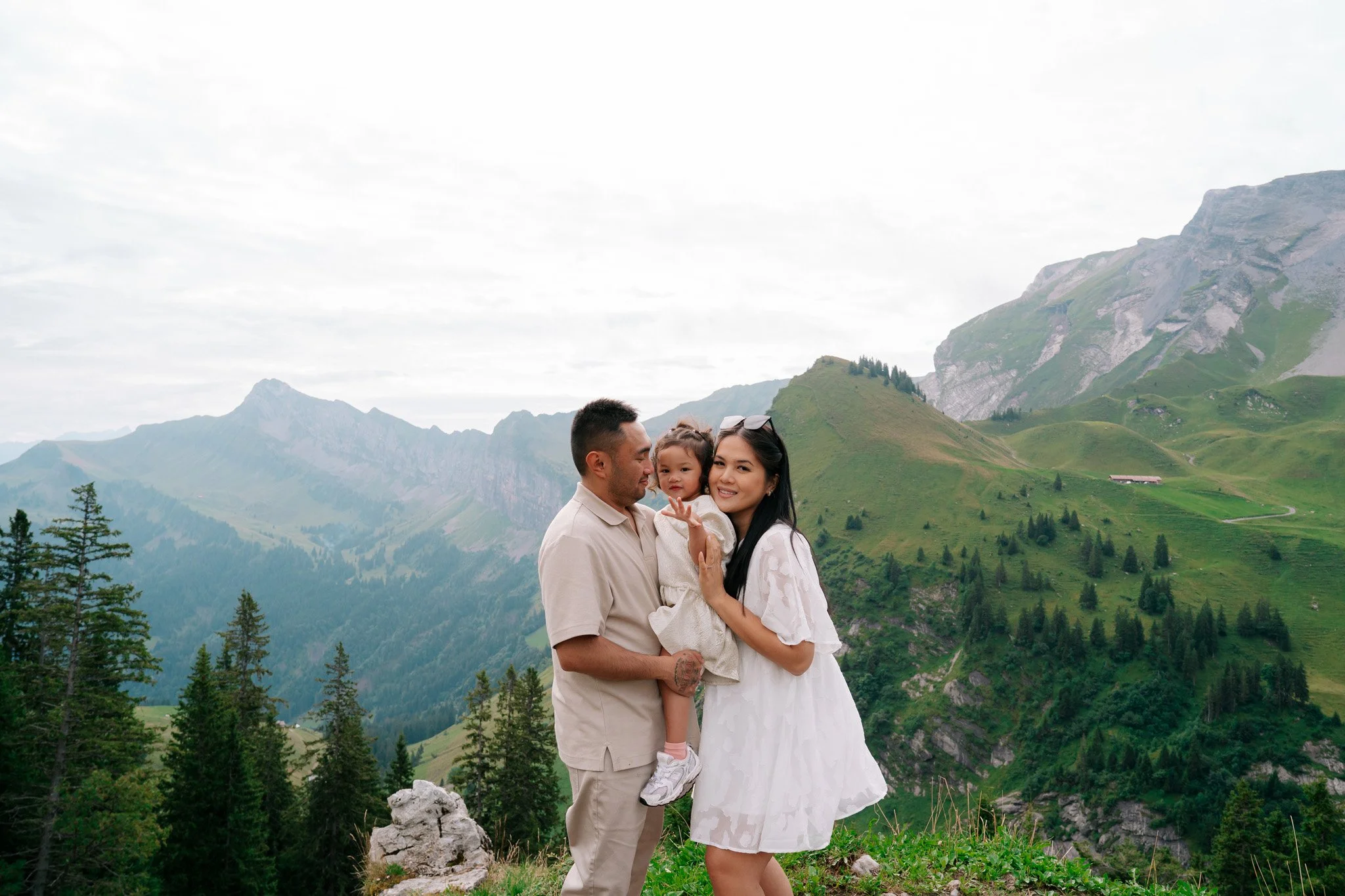 Family of three in mountain landscape, the man holding a young girl, woman smiling in white dress with mountains and trees in background. Family photoshoot in Switzerland.