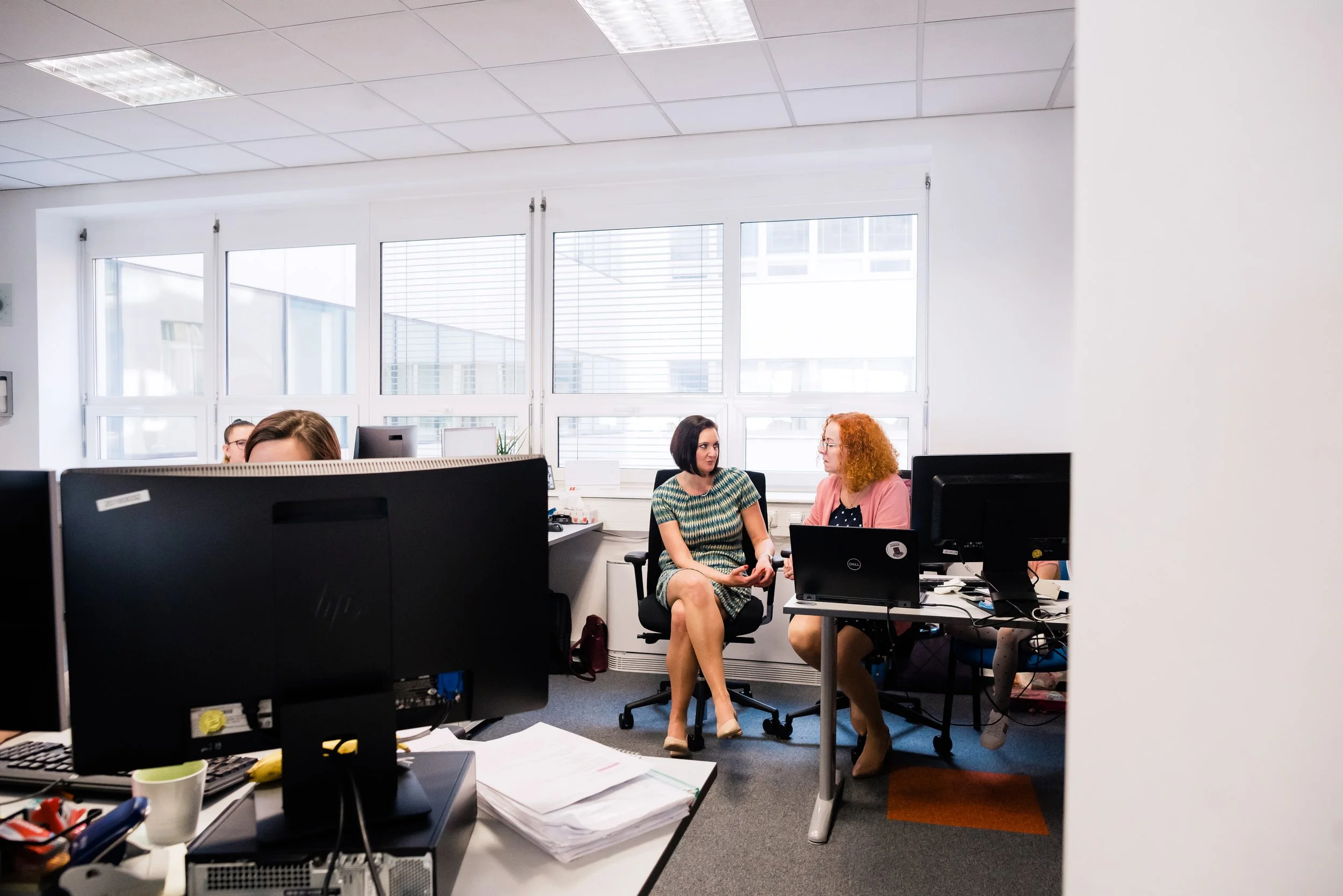 Two women having a conversation in a modern office, with computer monitors and desks in the foreground and windows in the background. Modern light and airy corporate company employer branding photography in Switzerland.