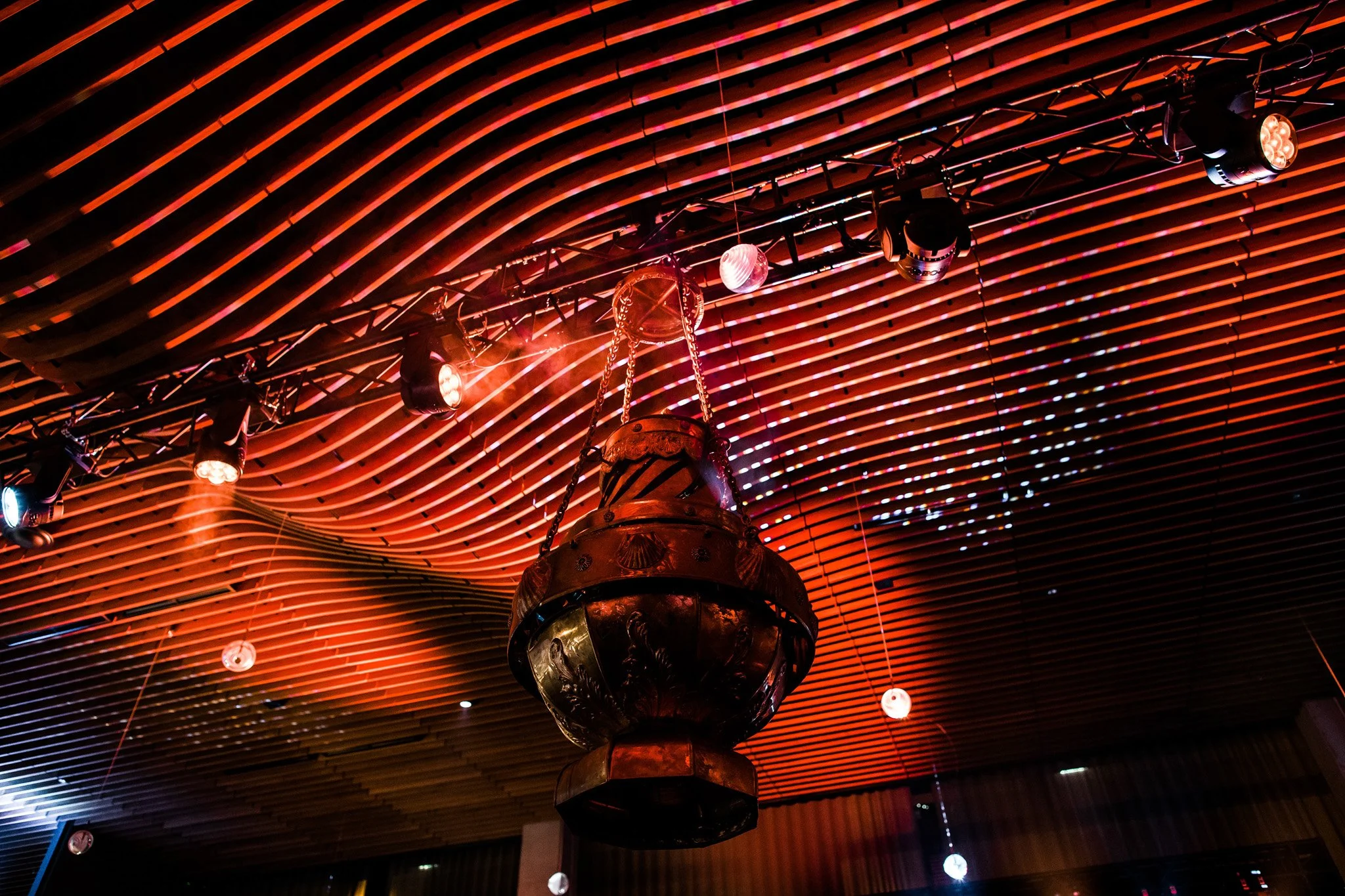 A large, ornate metal hanging lamp suspended from a curved wooden slatted ceiling with colorful lighting. Concert photography in Zürich, Zug, Bern, Lucerne, Basel, St. Gallen and across Switzerland.