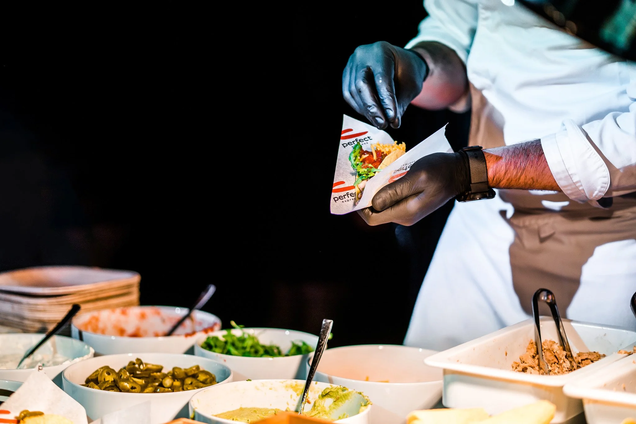 Chef wearing black gloves preparing a taco with various ingredients on a buffet table. Catering, culinary and food photography in Zürich, Zug, Bern, Lucerne, Basel, St. Gallen and across Switzerland.