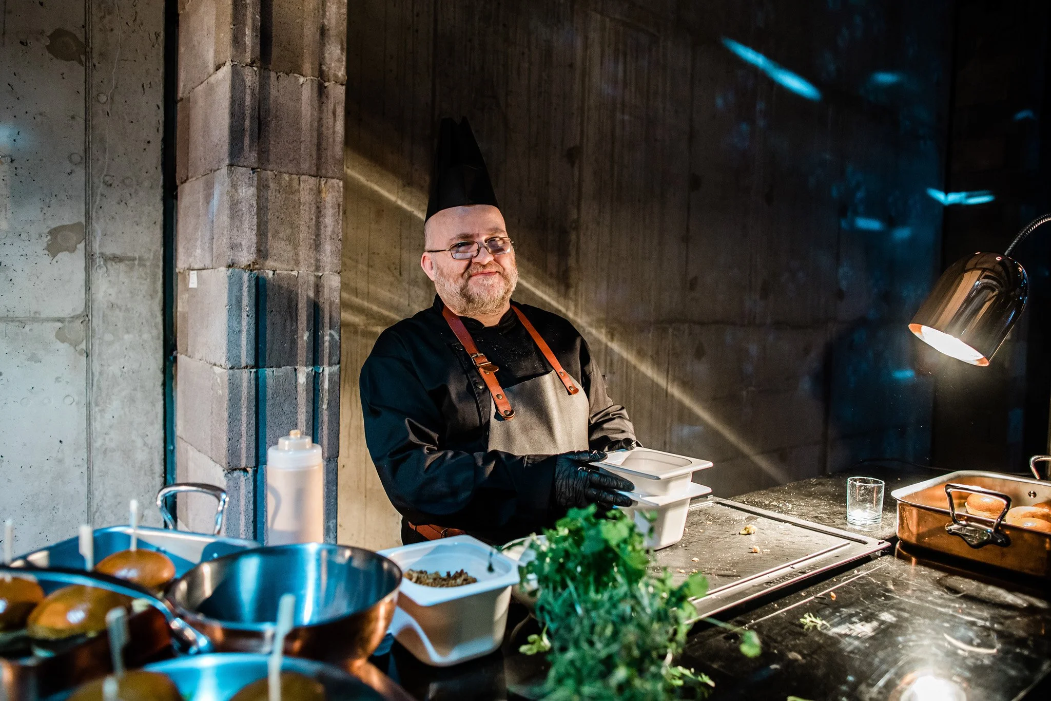 A chef with glasses and a beard, wearing a black chef's hat, apron, and gloves, standing in a professional kitchen, holding a container and smiling at the camera. Catering and food photography in Zürich, Zug, Bern, Lucerne and across Switzerland.