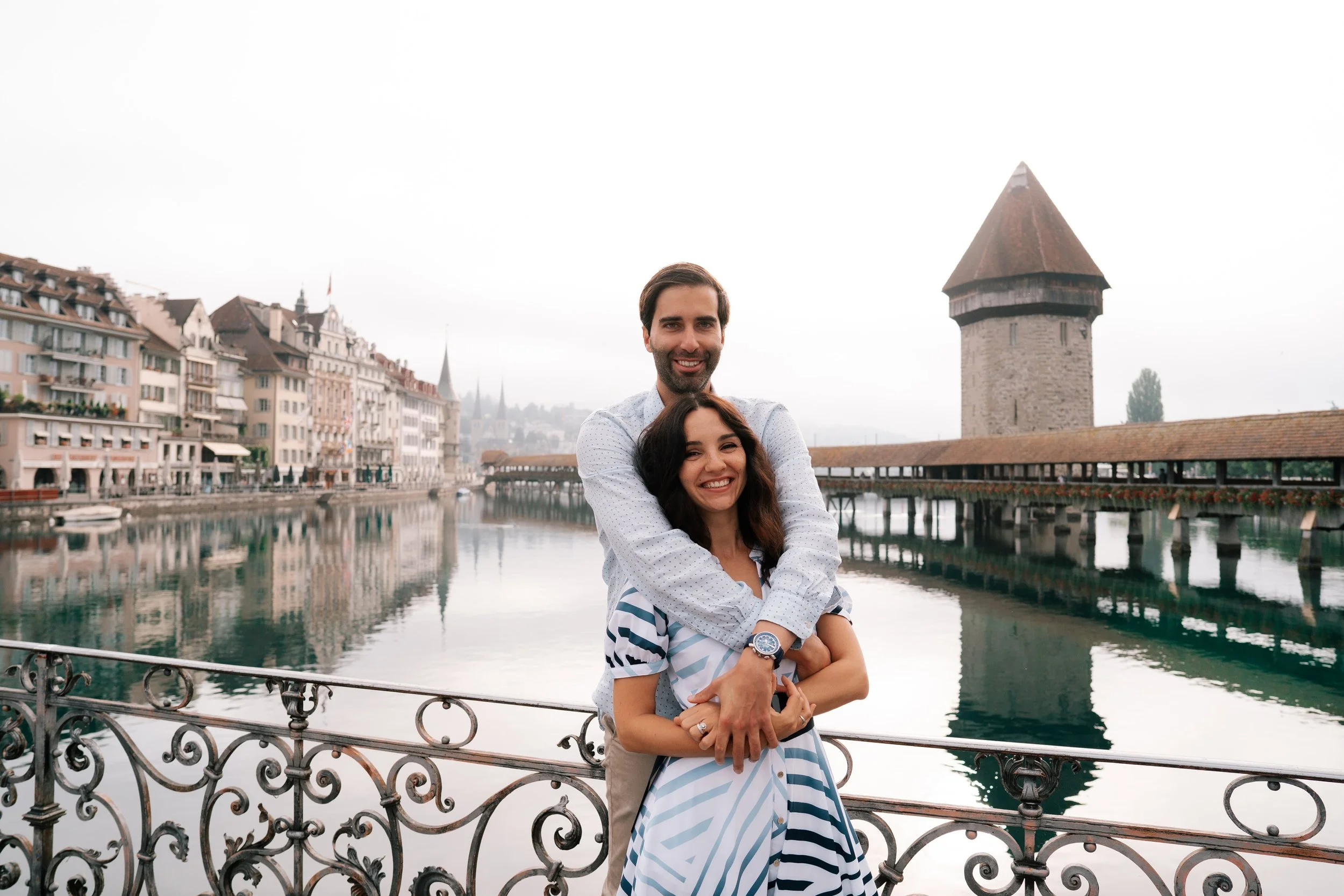 Smiling couple standing by a river in Lucerne with historic buildings and a medieval tower in the background.
