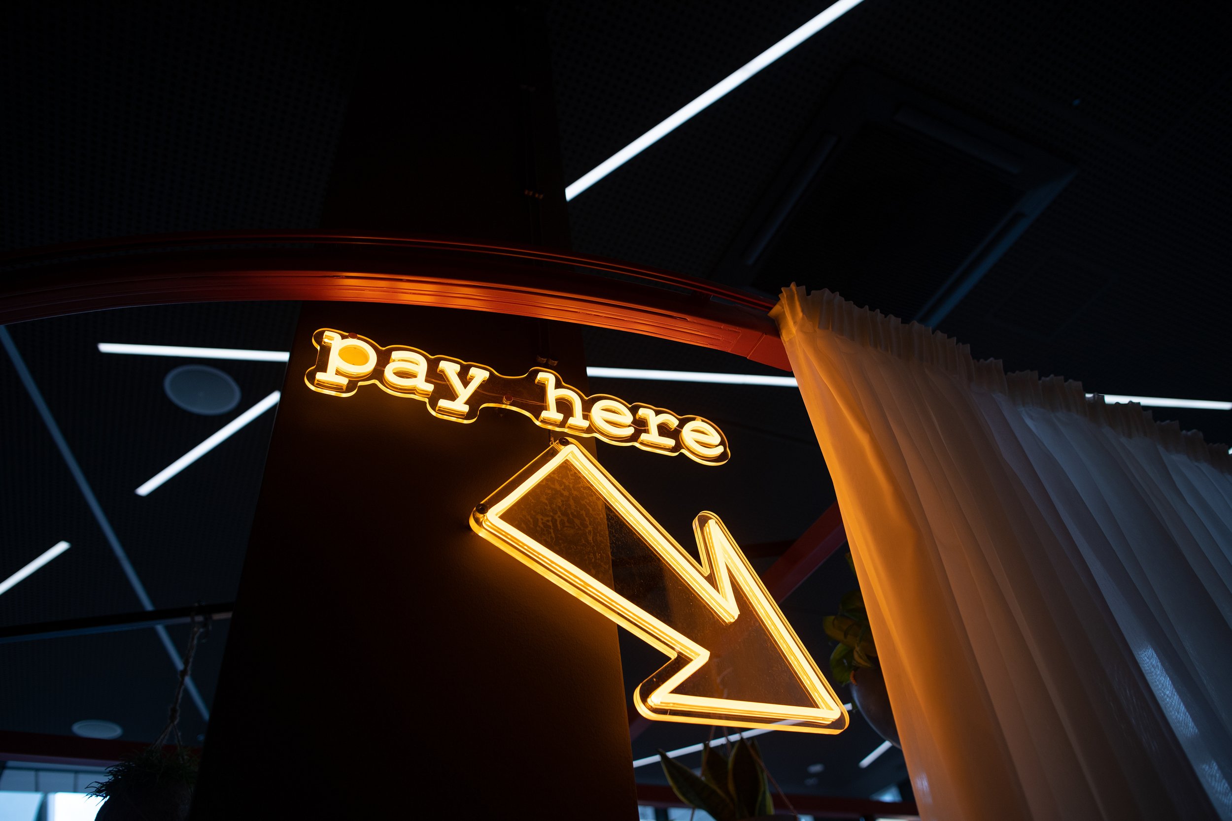 Neon sign that reads 'pay here' with a downward pointing arrow, illuminated in yellow, next to a white curtain in a modern indoor space. Restaurant interior photography in Zürich, Zug, Bern, Lucerne, Basel, St. Gallen and across Switzerland.