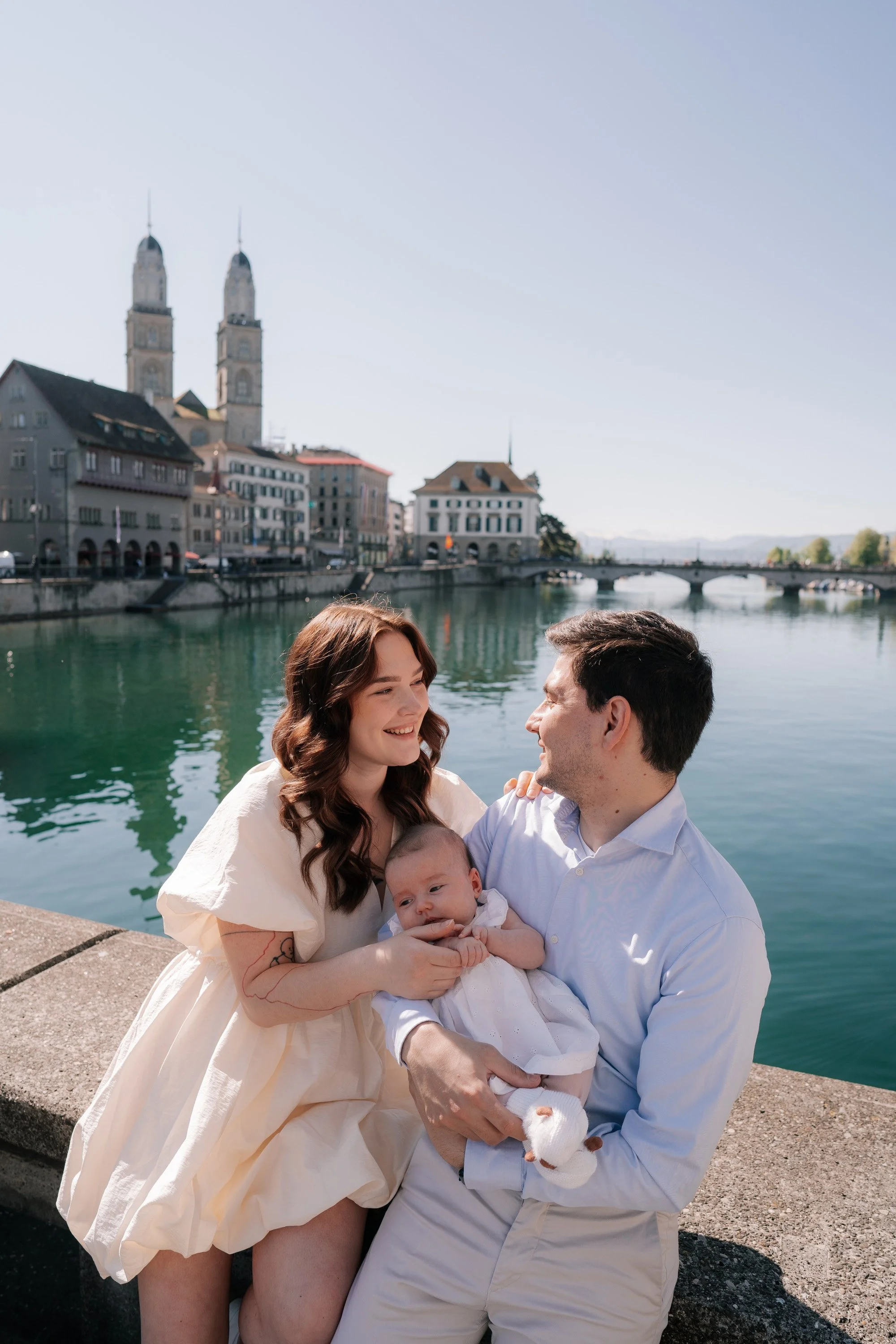 A family of three, including a woman, a man, and a baby, sitting by a river with historic buildings and church towers in the background on a sunny day in Zurich Switzerland.