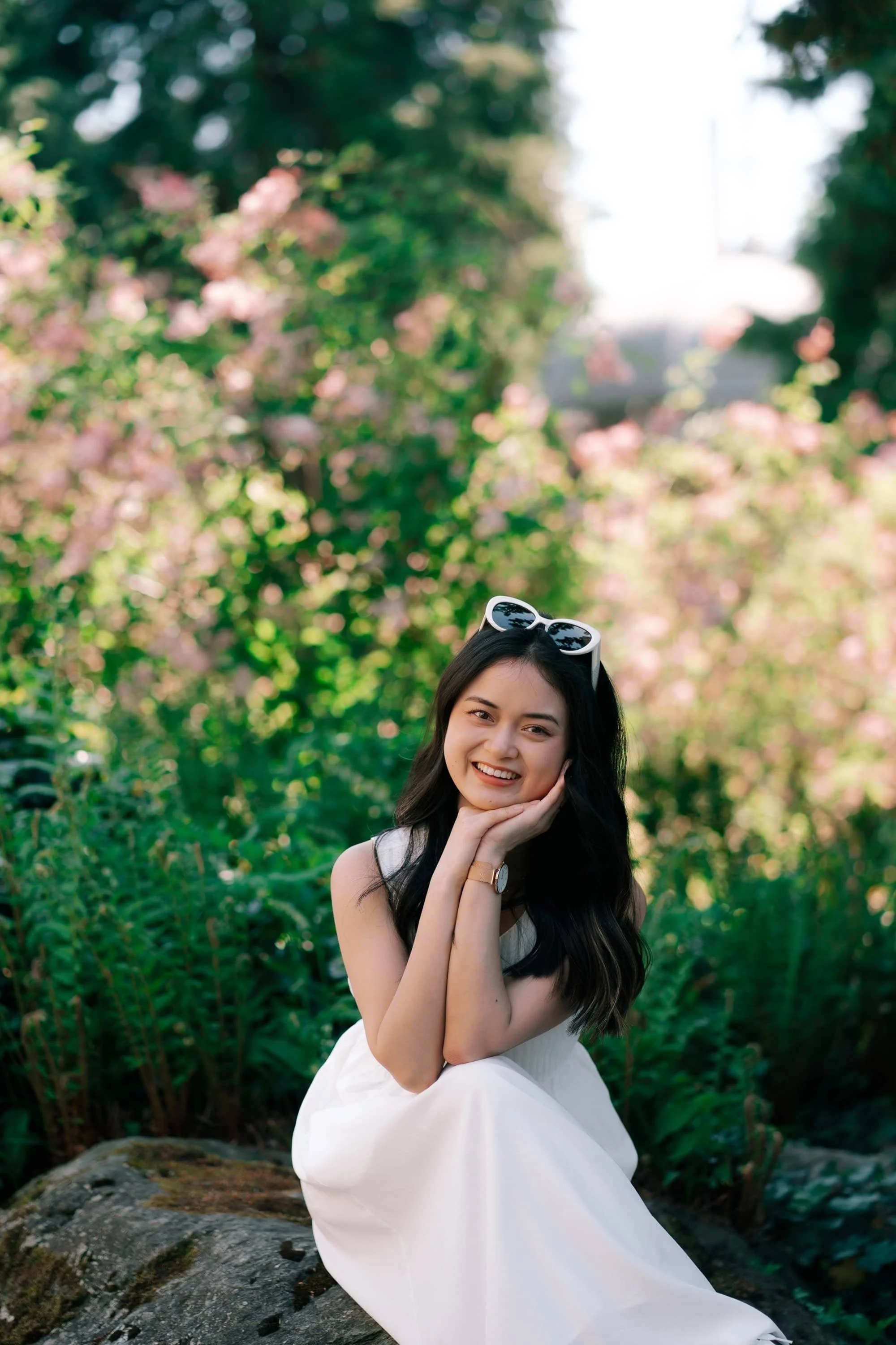 A young woman with long dark hair, wearing a white dress and sunglasses on her head, smiling and sitting on a rock in a lush green garden with pink flowers and trees. Villa Patumbah in Zurich.