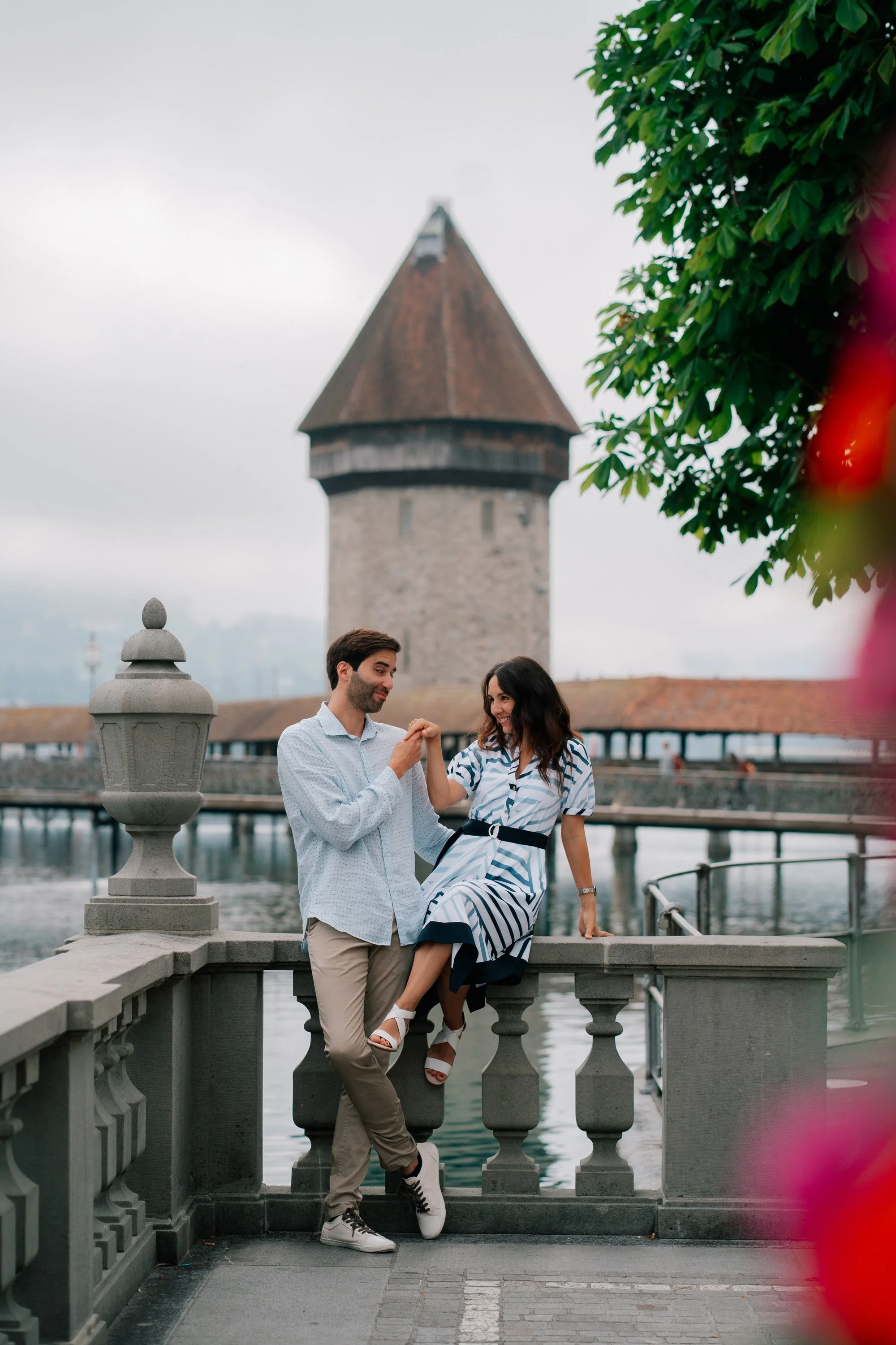 A man and woman holding hands and smiling by a lake with a castle tower in the background, on a cloudy day in Lucerne Switzerland.