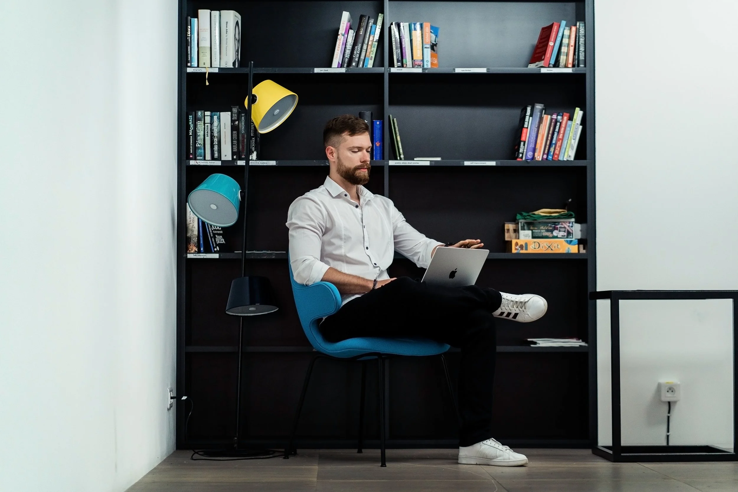 A man sitting on a blue office chair with his legs crossed, working on a silver laptop, seated in front of a black bookshelf filled with books and colorful desk lamps in a modern office space. Business headshot, personal branding Switzerland.