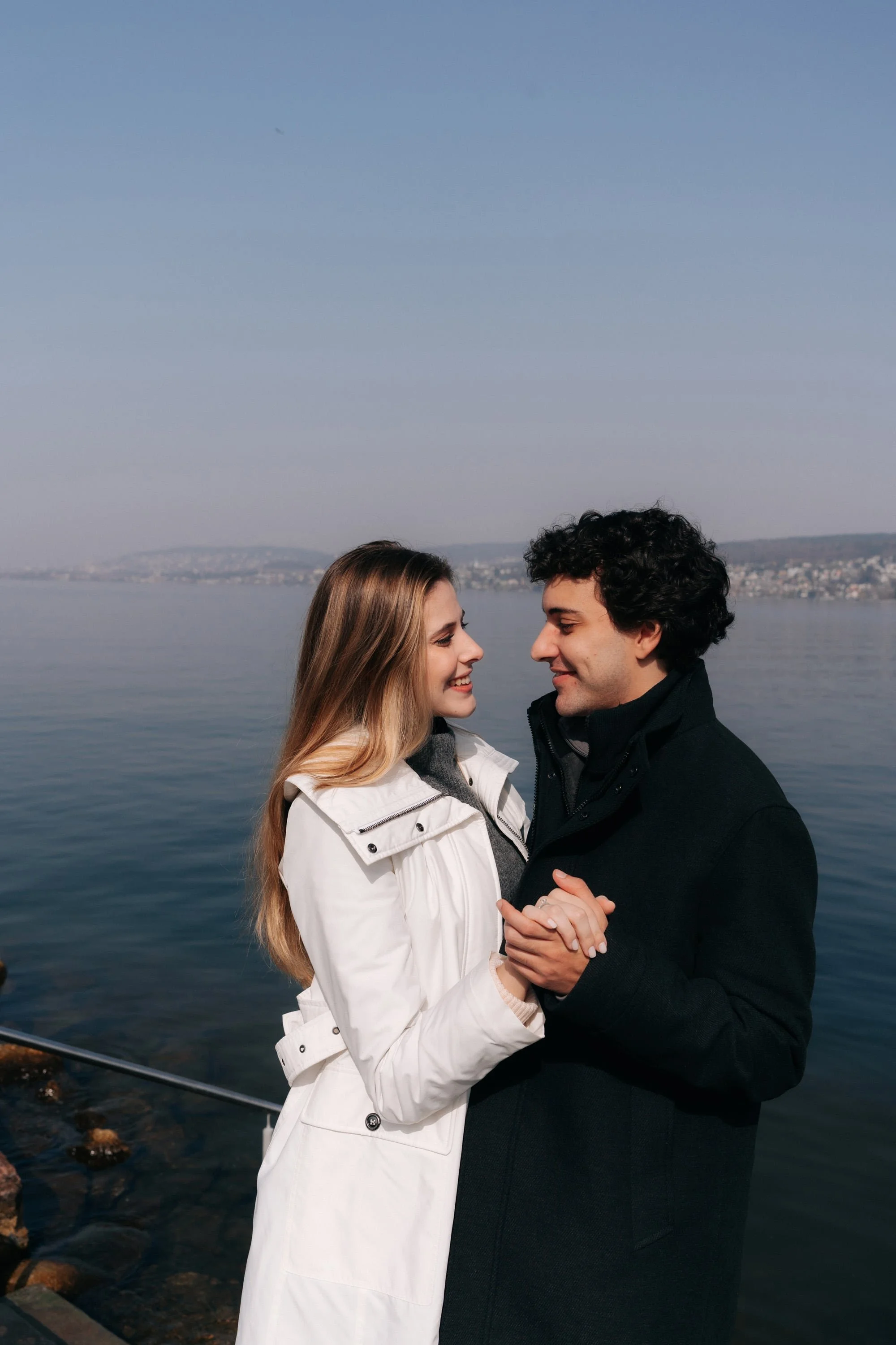 A young couple stands close together by a body of water, smiling and holding hands, with a cityscape and distant hills under a clear blue sky in the background. They are celebrating their surprise proposal in Switzerland in Thalwil.
