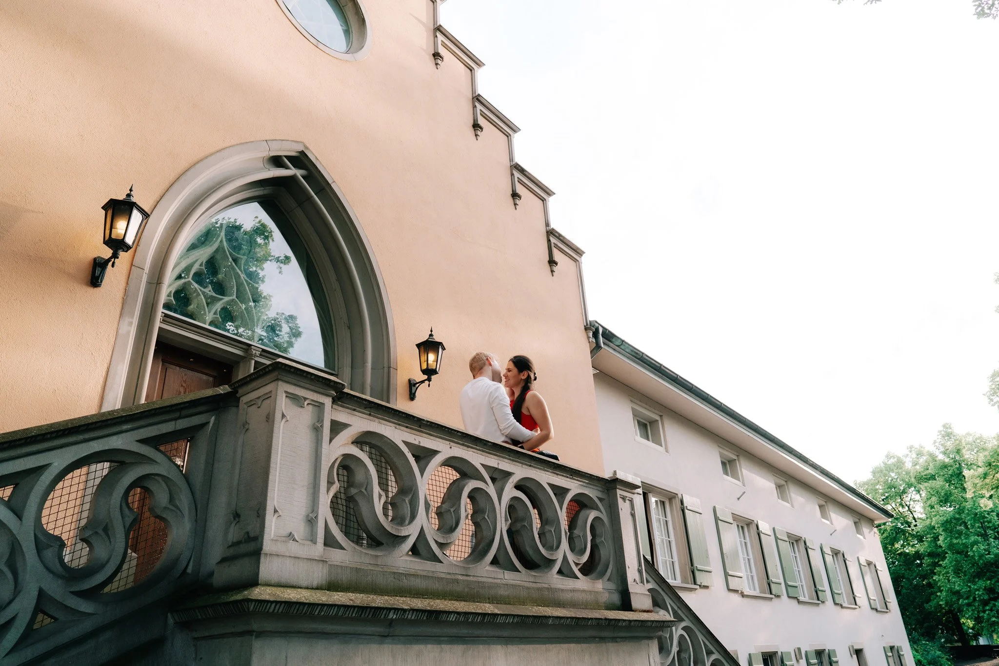 A couple standing on a balcony of a large house in Zurich Switzerland, smiling at each other, with the house's peach-colored exterior wall and ornate railing visible. The house has multiple windows with shutters and wall-mounted lanterns.