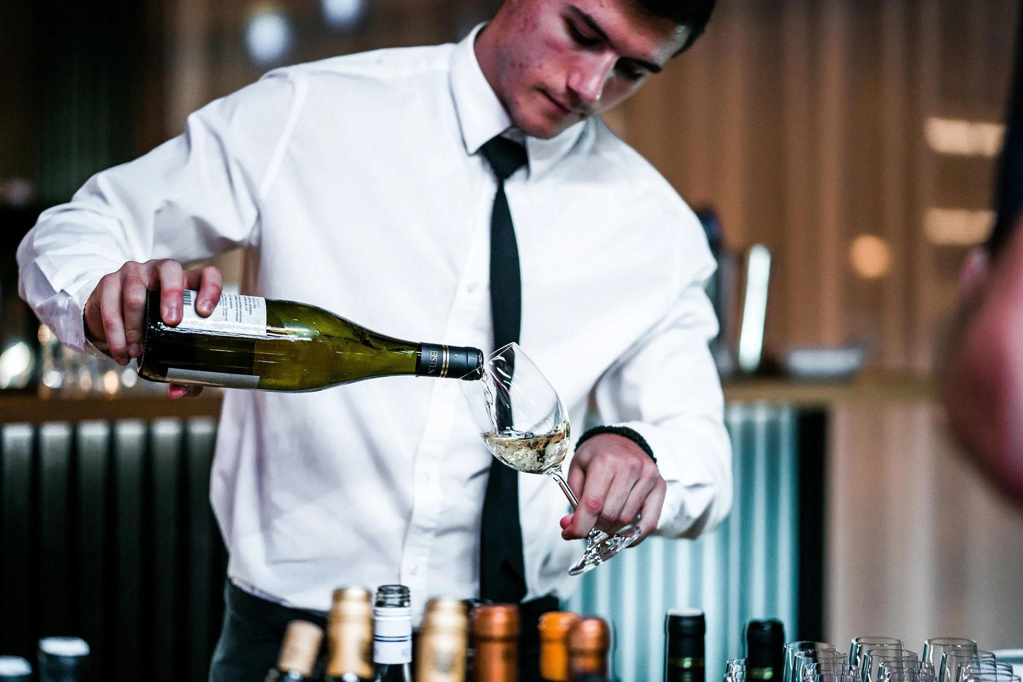 A man in a white shirt and black tie pouring white wine into a glass at a wine tasting event. Catering, culinary and food photography in Zürich, Zug, Bern, Lucerne, Basel, St. Gallen and across Switzerland.