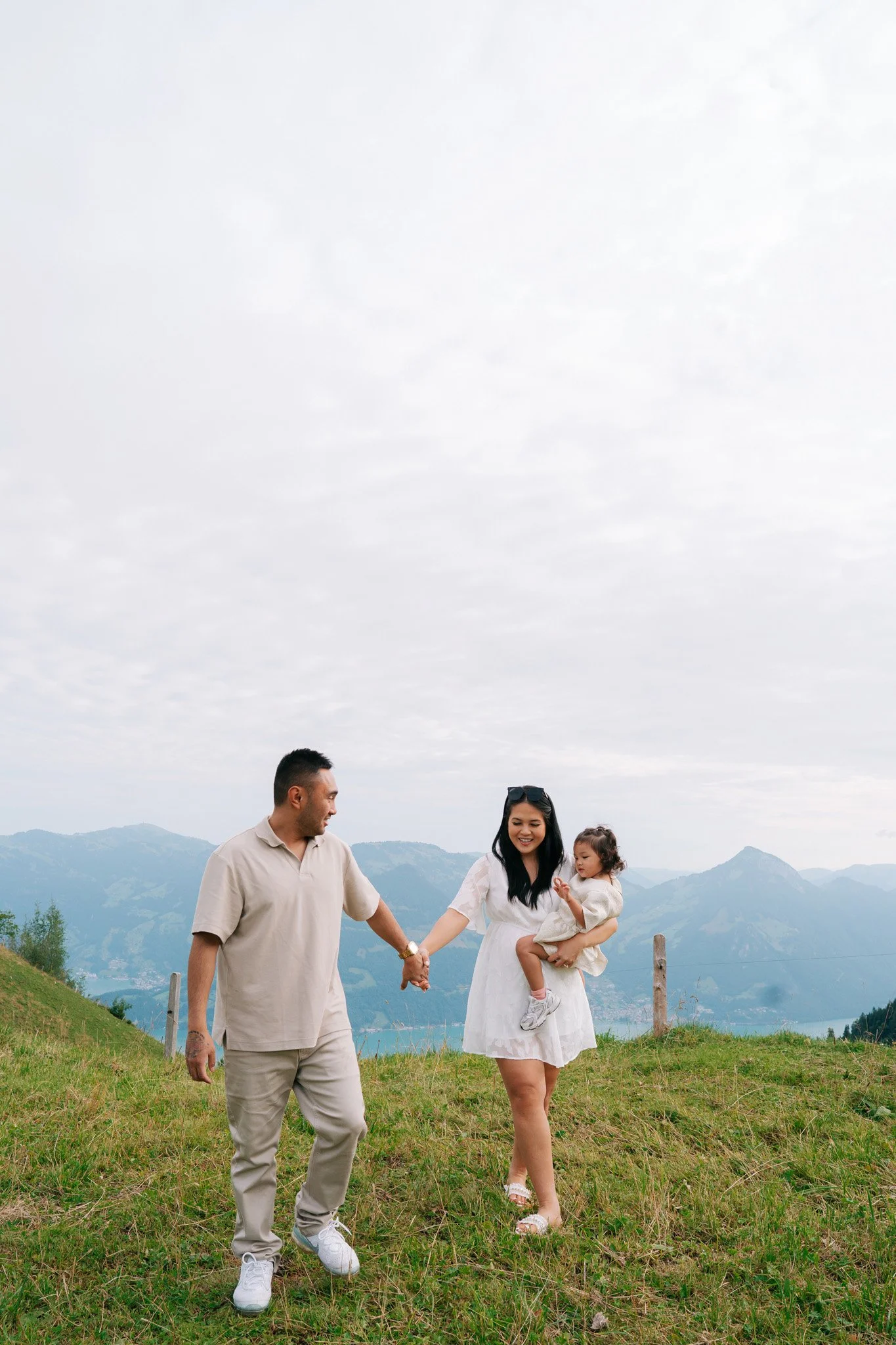A family of three walking outdoors in a grassy field with mountains in the background. The man is holding the woman's hand, and the woman is holding a young girl. The sky is partly cloudy. Family photoshoot in Switzerland.