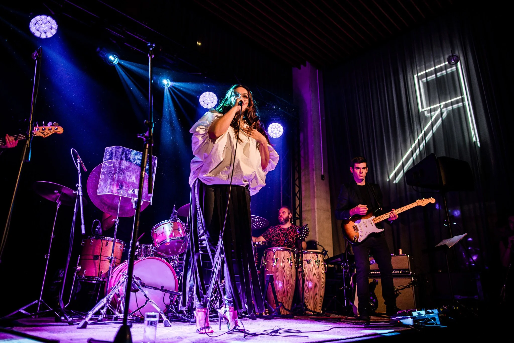 Band performing on stage with vocalist holding a microphone, drummer, percussionist, and guitarist, colorful stage lighting, neon decor, and musical equipment. Concert photographer in Zürich, Zug, Bern, Lucerne, Basel, St. Gallen and Switzerland.