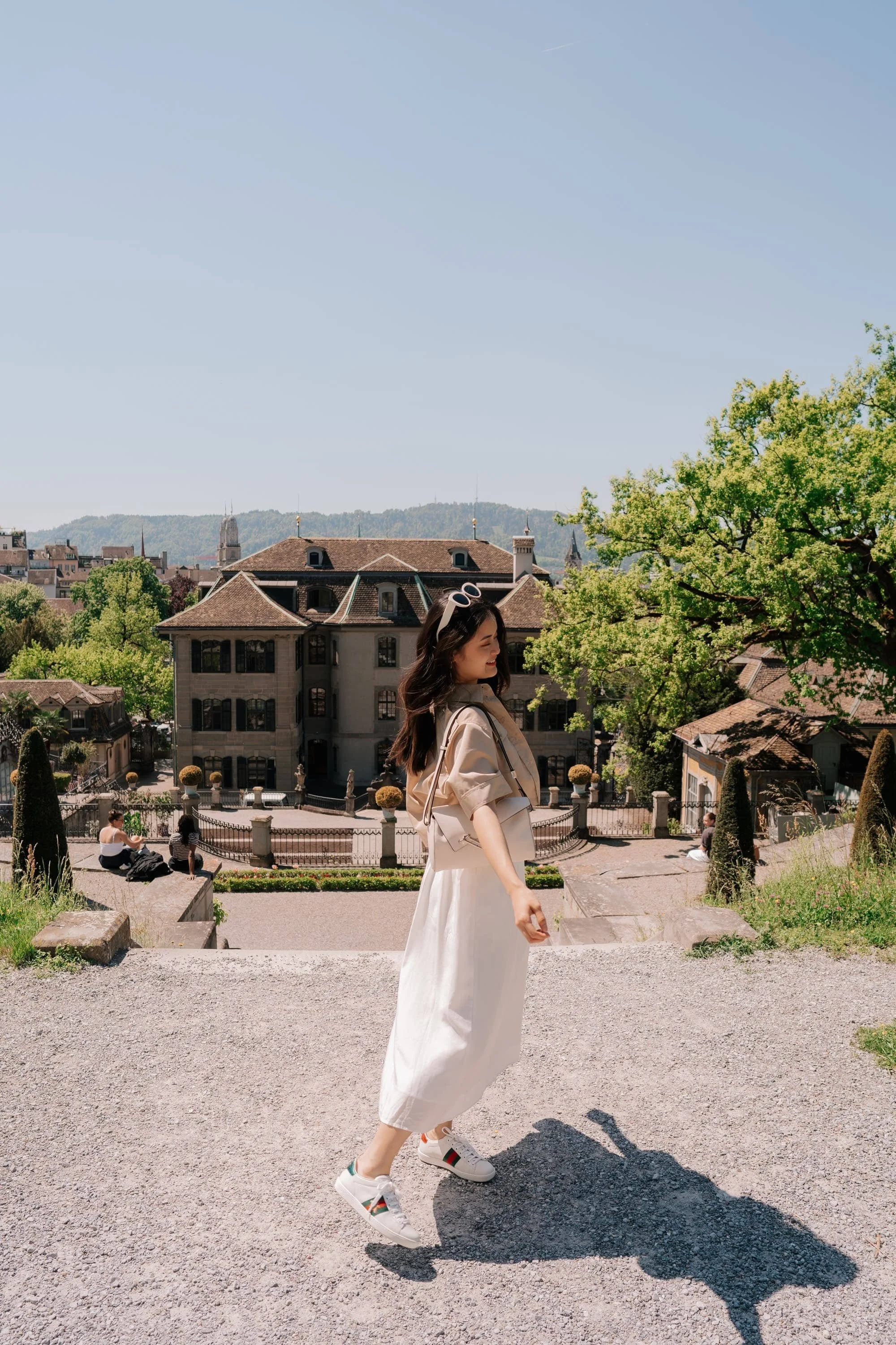 A woman in a beige jacket and white skirt walks outdoors on a sunny day with a city and hills in the background. She wears sunglasses on her head and sneakers. Gardens in Zurich.