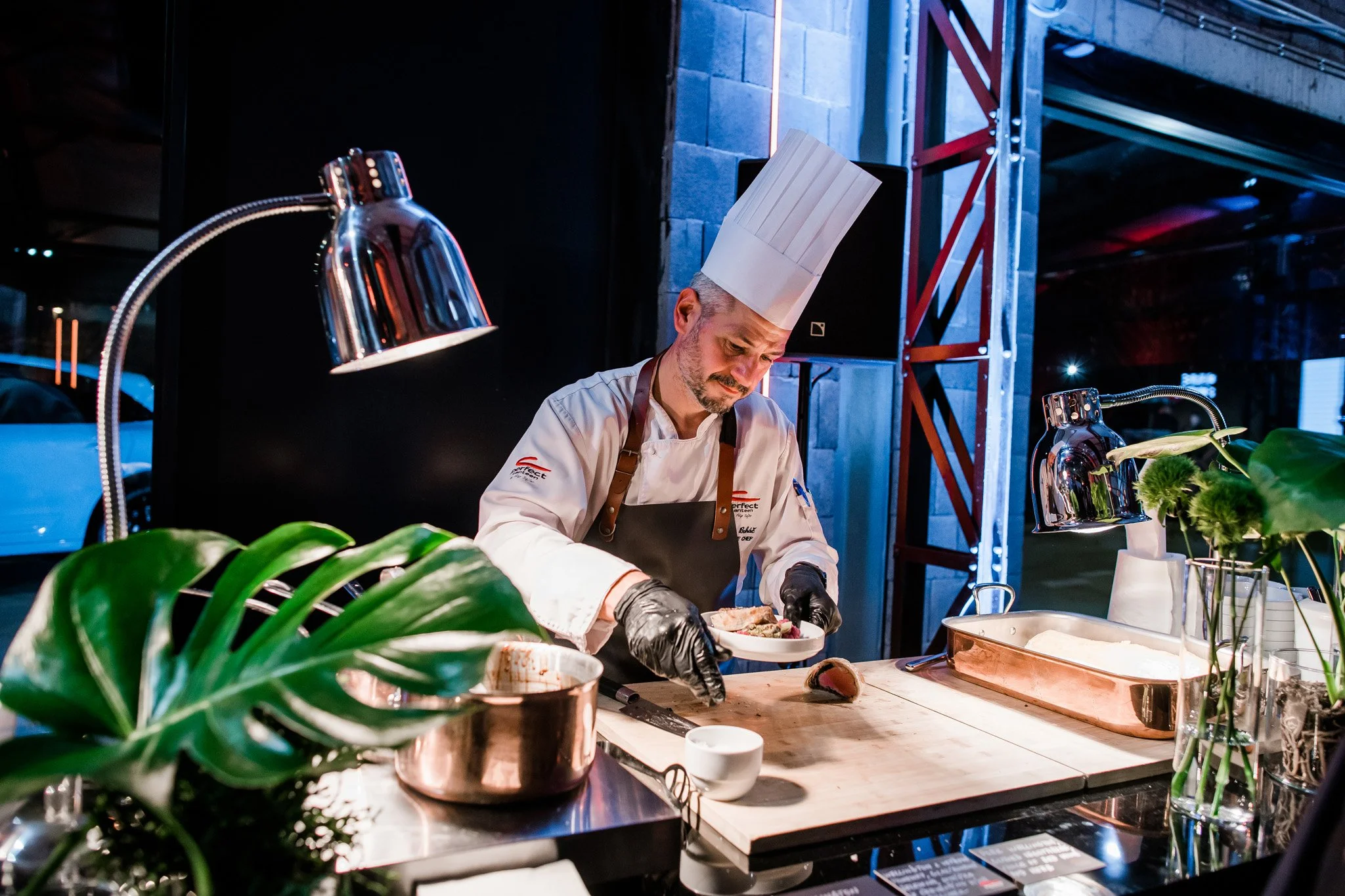 A chef wearing a tall white hat, black gloves, and a white apron is preparing food in a modern kitchen with industrial decor. The chef is slicing a piece of meat on a wooden cutting board, with a copper pot and some green plants visible on the counte