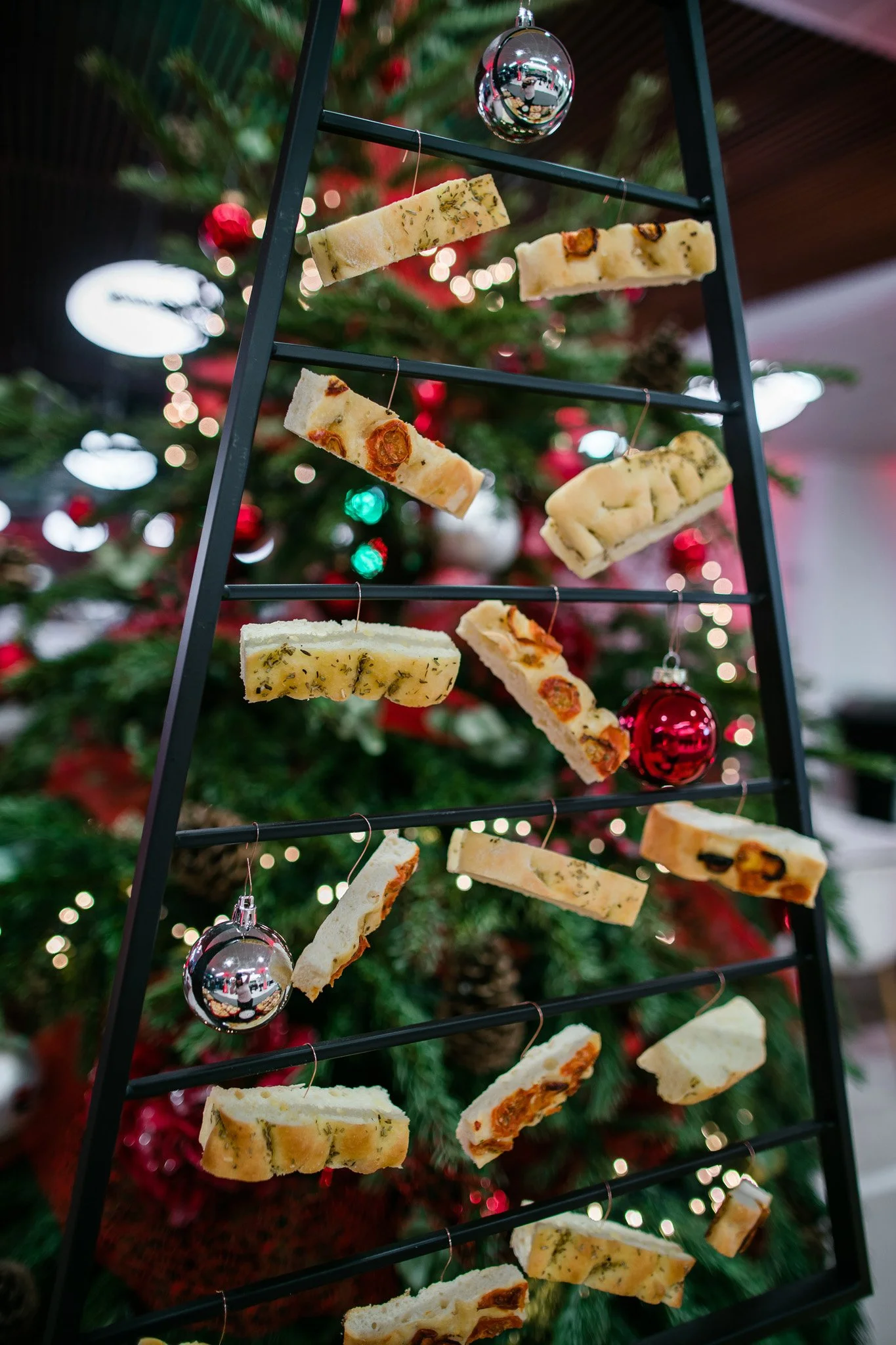 Christmas tree decorated with red and silver baubles in a festive setting, with a black metal frame display holding slices of assorted cheese, some with herbs, spices, or fruit. Catering, culinary and food photography in Switzerland.