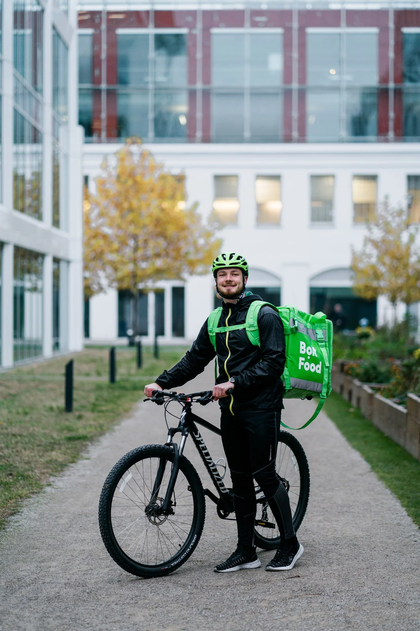 A man in black athletic clothing and helmet smiles while standing with a black mountain bike. He carries a large green insulated food delivery bag labeled 'Bon Food' on his back. Advertising campaign photographer for corporate and startup Switzerland