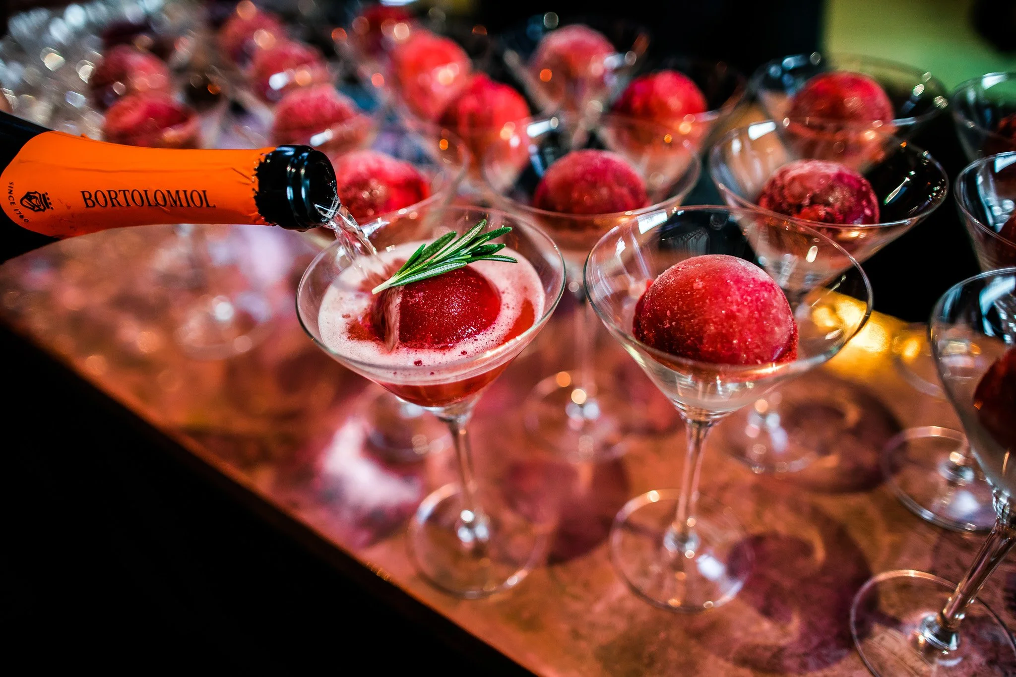 Champagne being poured into a cocktail glass with a raspberry garnish, surrounded by other raspberry cocktails on a table. Catering and food photography in Zürich, Zug, Bern, Lucerne, Basel, St. Gallen and across Switzerland.