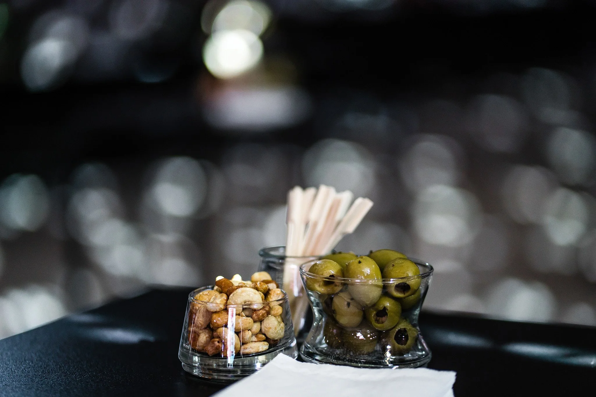 Small glass bowls containing green olives, mixed nuts, and toothpicks on a black surface with a blurry background. Catering and food photography in Zürich, Zug, Bern, Lucerne, Basel, St. Gallen and across Switzerland.