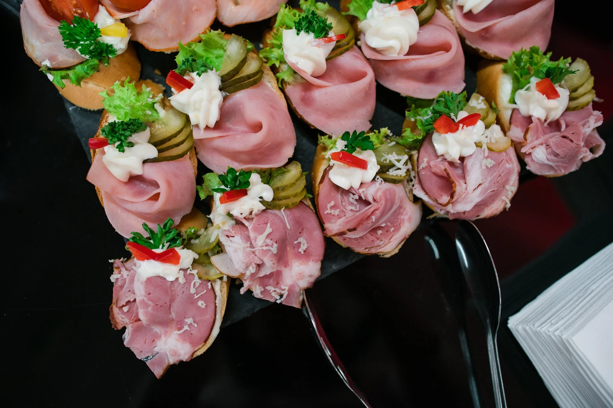 Assorted open-faced sandwiches with ham, lettuce, pickles, mayonnaise, parsley, and red bell pepper on small bread slices. Catering, culinary and food photography in Zürich, Zug, Bern, Lucerne, Basel, St. Gallen and across Switzerland.