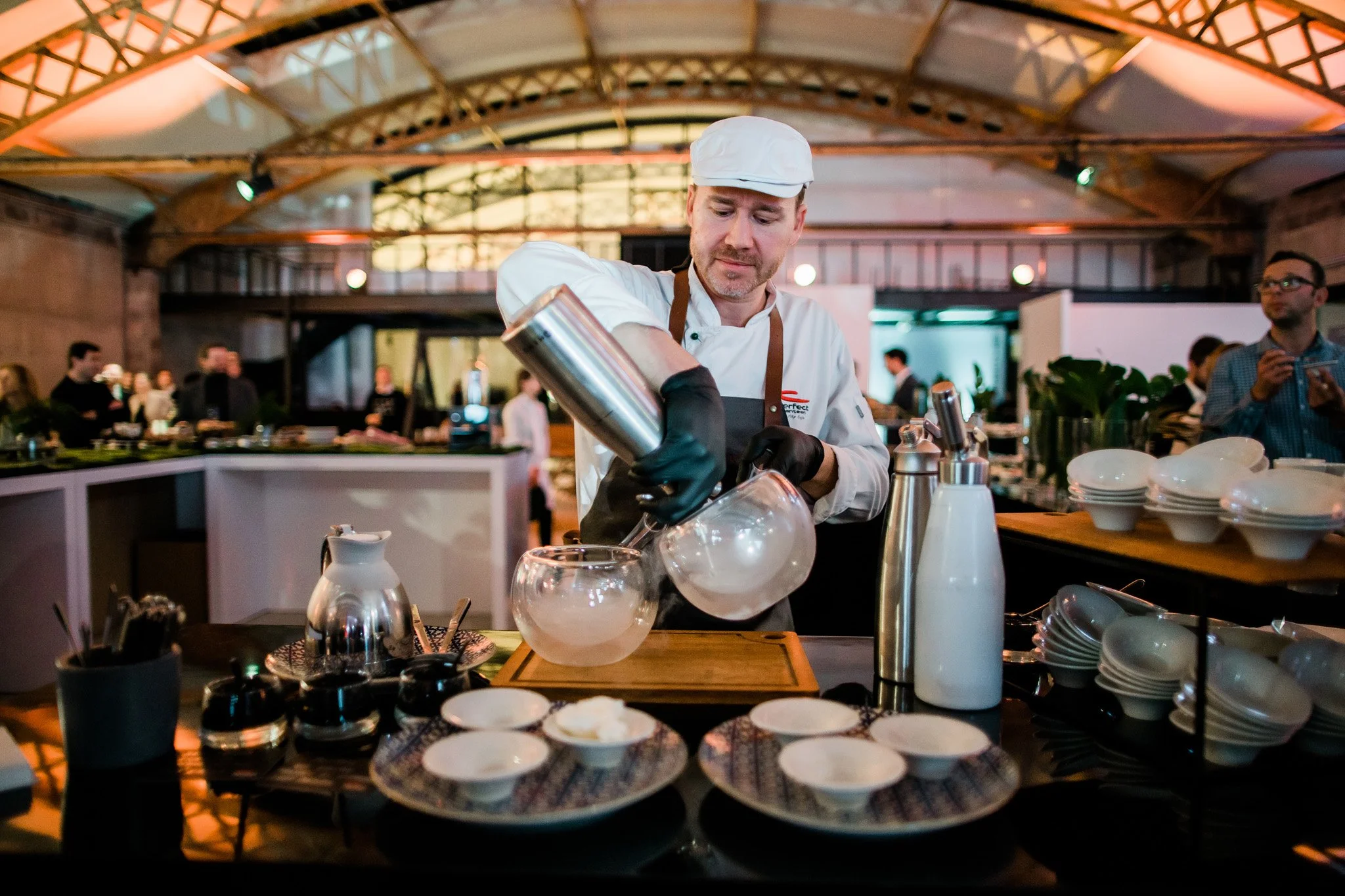 Chef in a white uniform and black gloves preparing a cocktail with dry ice in a glass bowl at a bar in a large, industrial-style venue with other people in the background. Catering and food photography in Zurich, Switzerland. 