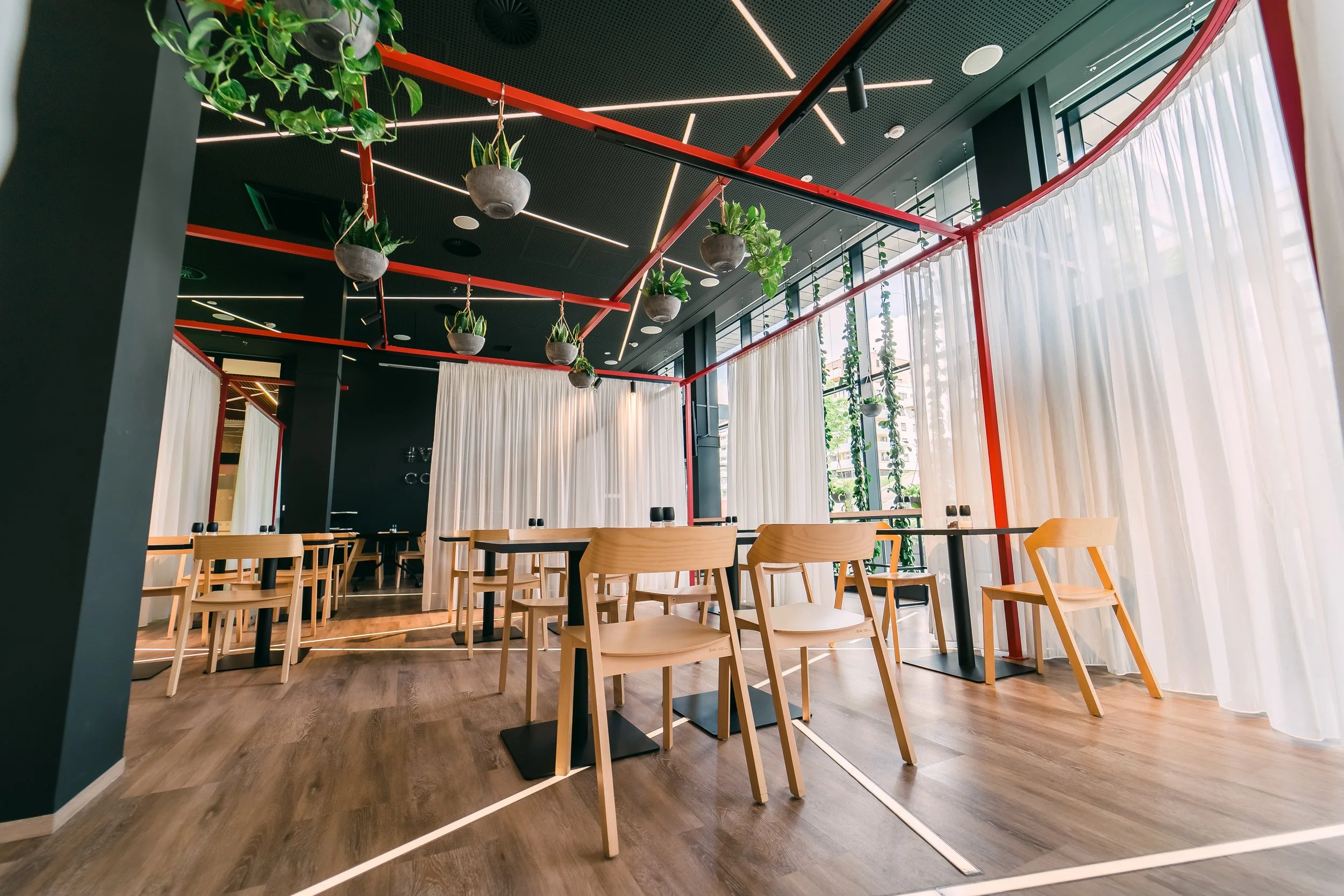 Modern restaurant with wooden chairs, long tables, hanging plants, white curtains, large windows, and decorative ceiling lighting. Restaurant interior photography in Zürich, Zug, Bern, Lucerne, Basel, St. Gallen and across Switzerland.