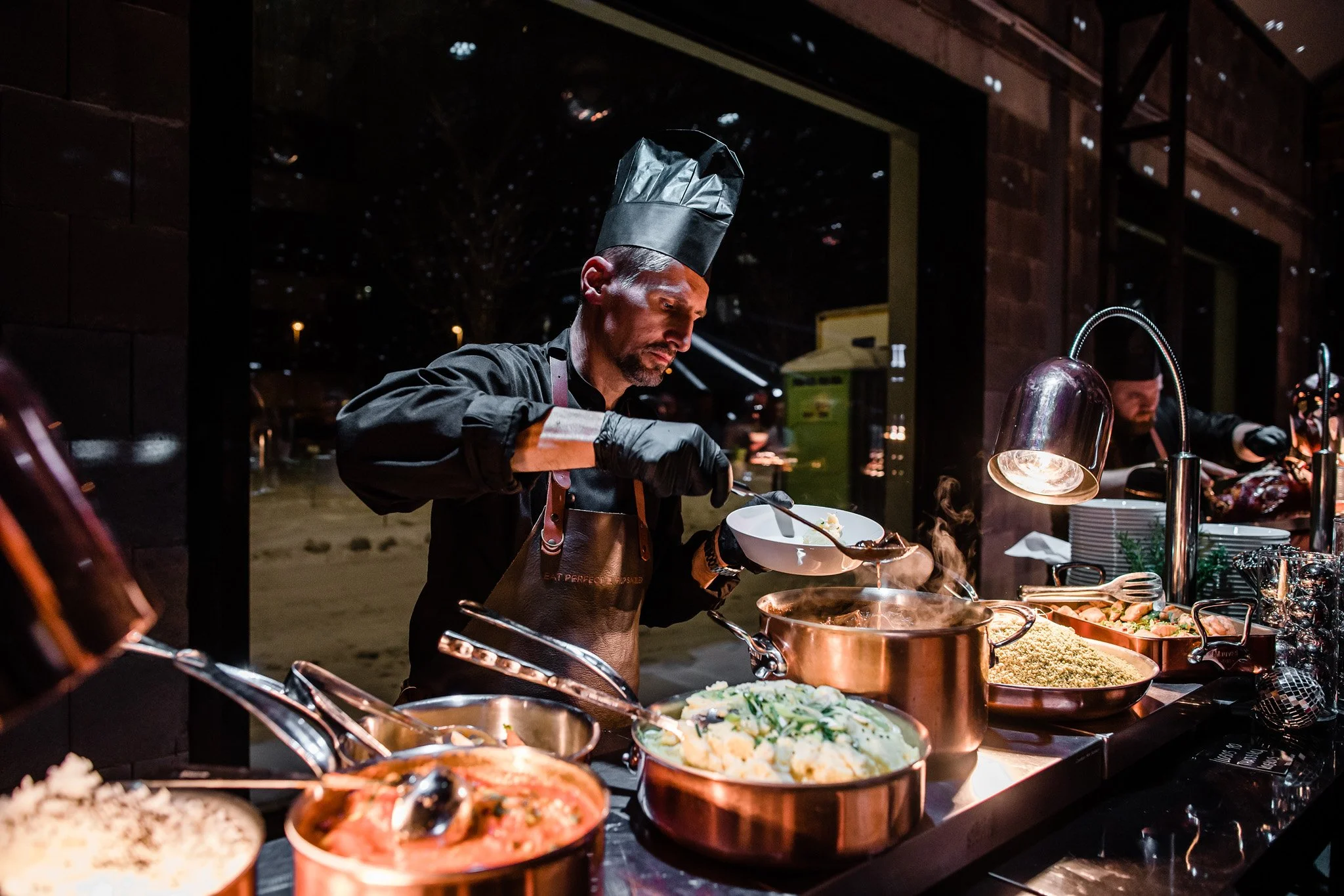 Chef in black uniform and hat serving food at buffet table with various dishes and hot lamps in a restaurant with large window at night. Catering and food photography in Zürich, Zug, Bern, Lucerne, Basel, St. Gallen and across Switzerland.