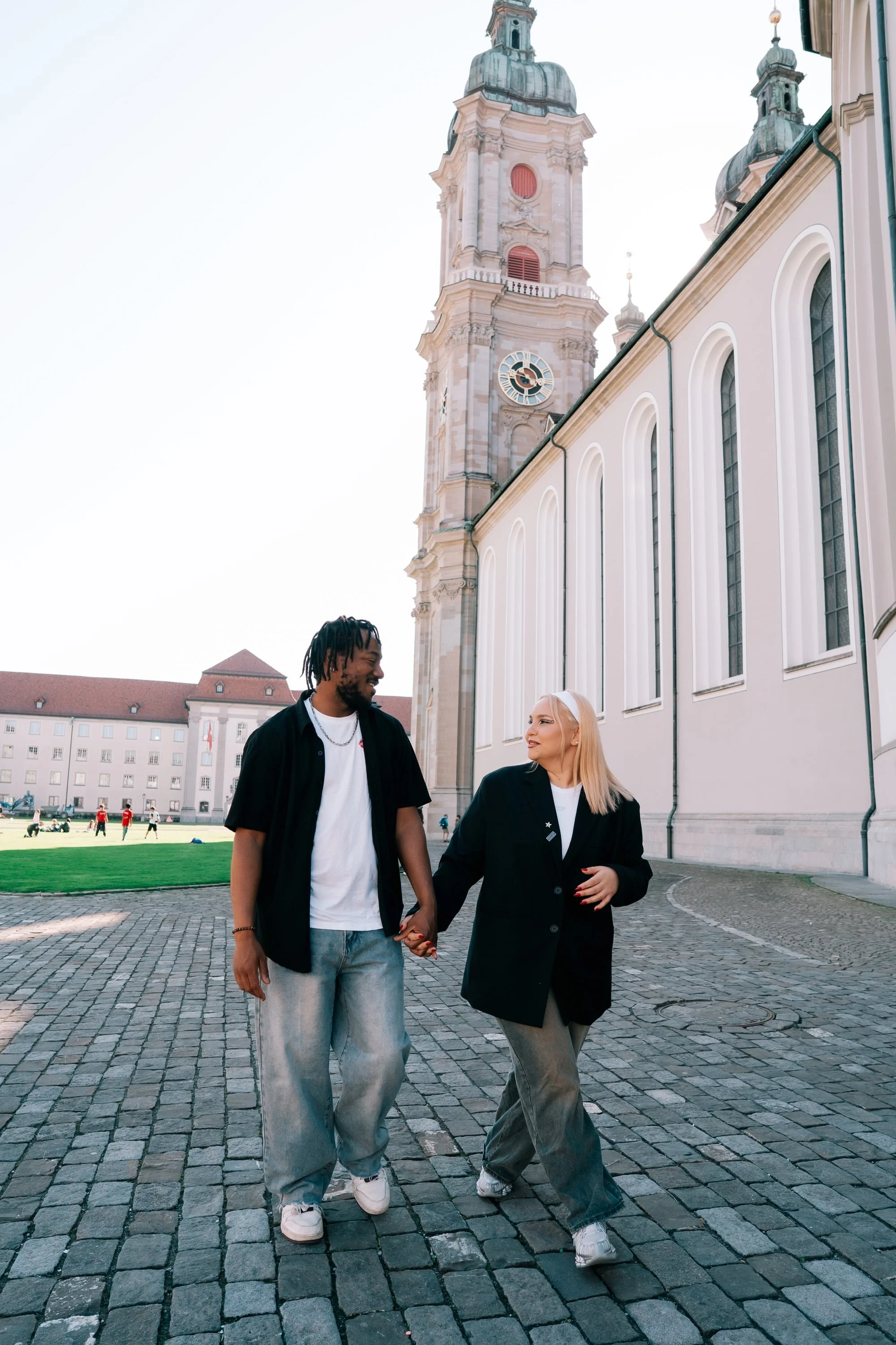 A couple walking hand in hand on a cobblestone street in front of a historic church in St Gallen Switzerland, with green grass and a building in the background.