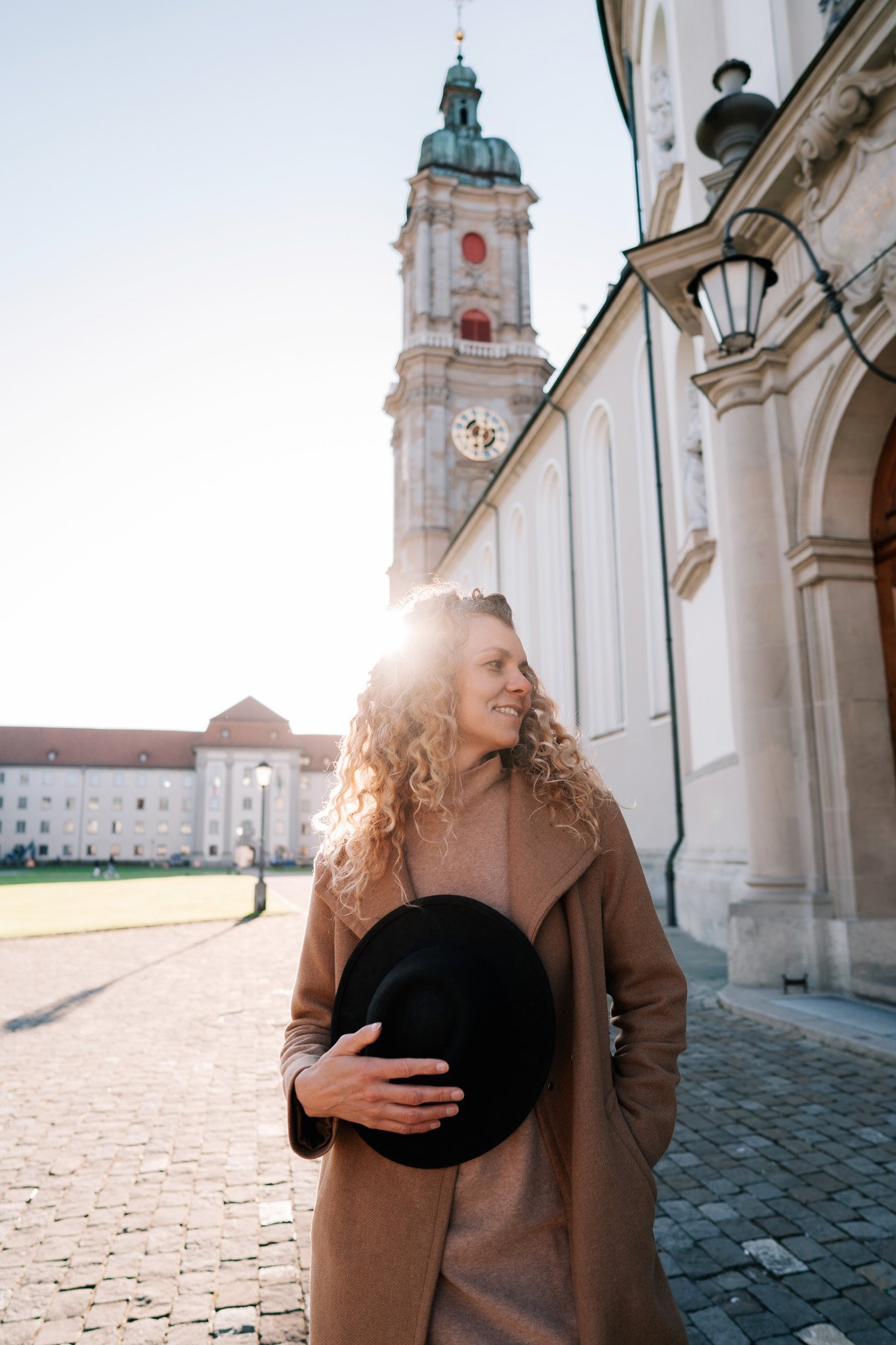 A woman with curly blonde hair standing outdoors in front of a historic building with a tall clock tower in St. Gallen Switzerland. She is smiling, holding a black hat with her right hand, wearing a beige coat.
