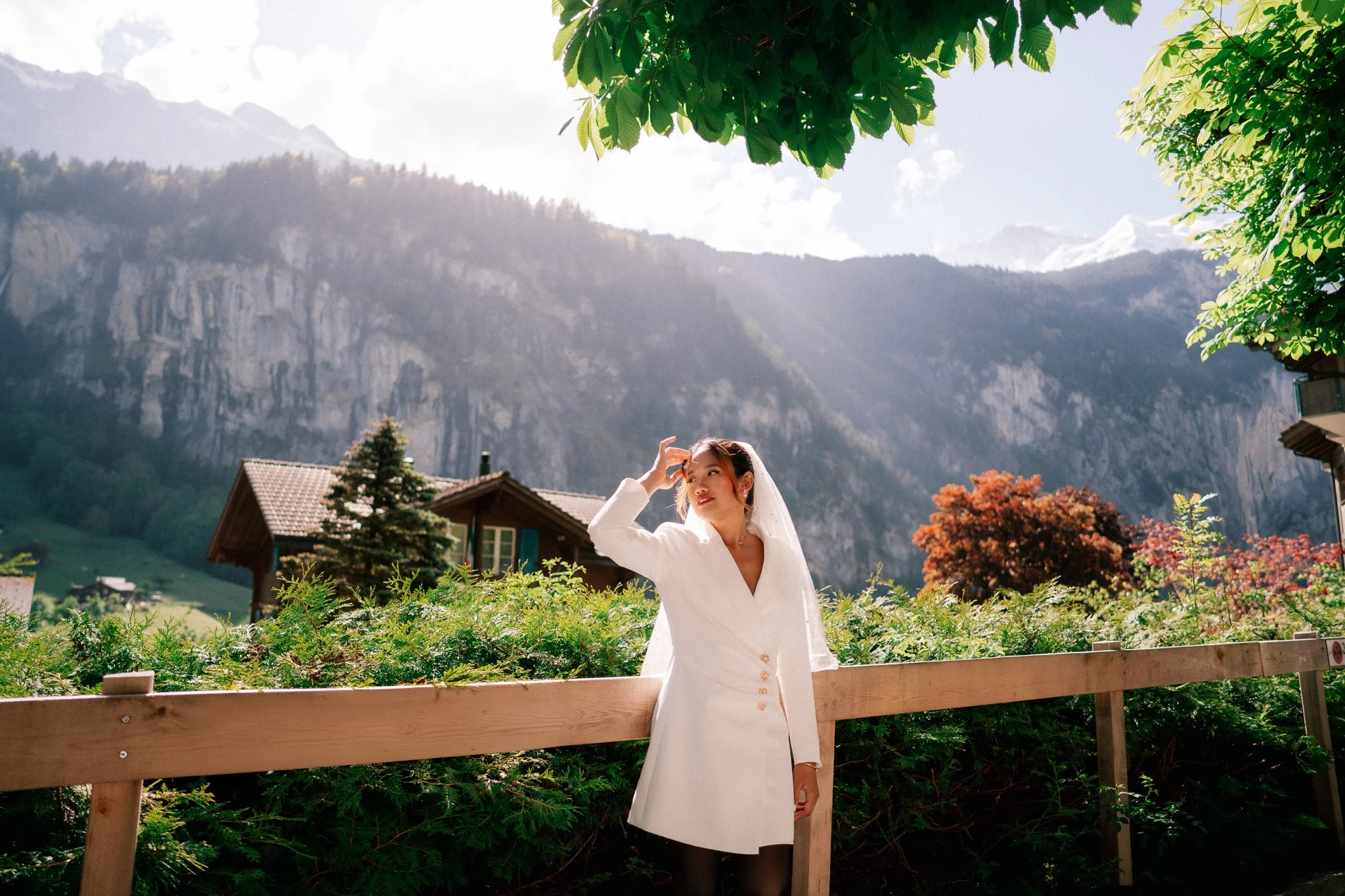 Wedding celebration photoshoot outdoors in front of a wooden fence, with mountains, trees, and a house in the background under a partly cloudy sky in Lauterbrunnen, Switzerland.