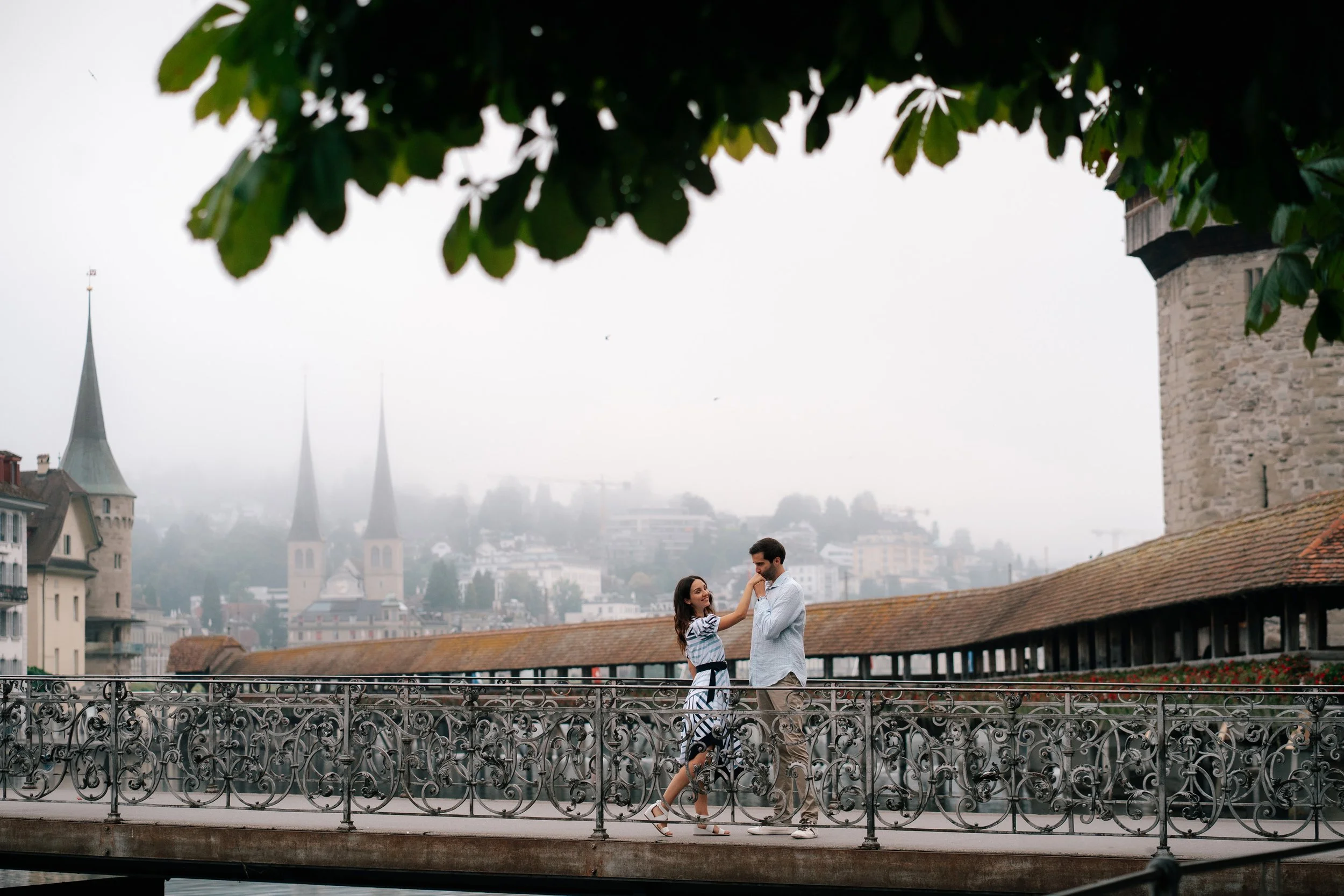 A couple on a decorative bridge in Lucerne Switzerland, with church spires and historic buildings in the background.