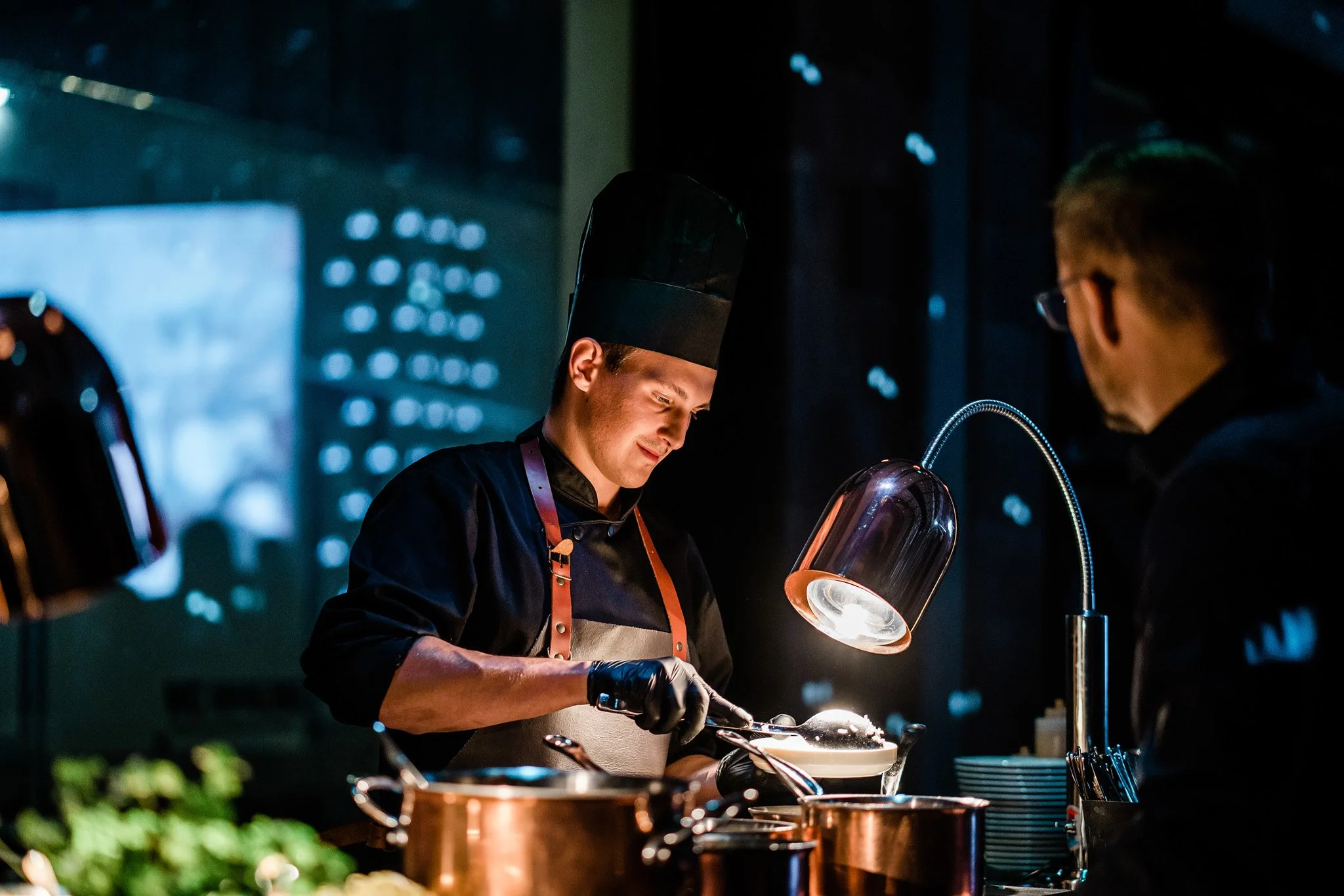 A chef in a tall black hat and apron preparing food in a professional kitchen under warm lighting, with another person observing. Catering and food photography in Zürich, Zug, Bern, Lucerne, Basel, St. Gallen and across Switzerland.
