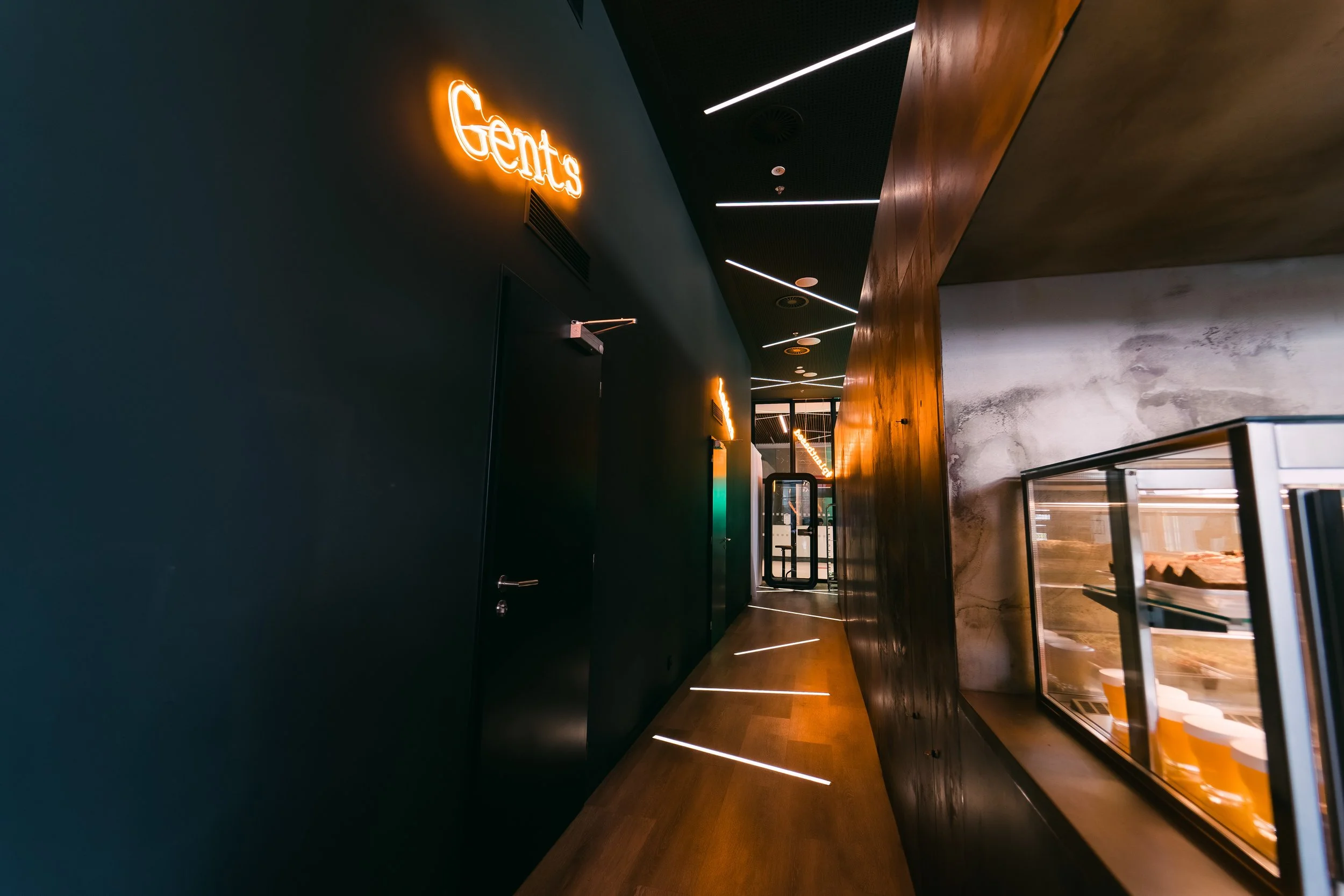 Modern hallway with emerald green wall, illuminated 'Gents' sign, wooden flooring, and glass display case with baked goods. Restaurant interior photography in Zürich, Zug, Bern, Lucerne, Basel, St. Gallen and across Switzerland.