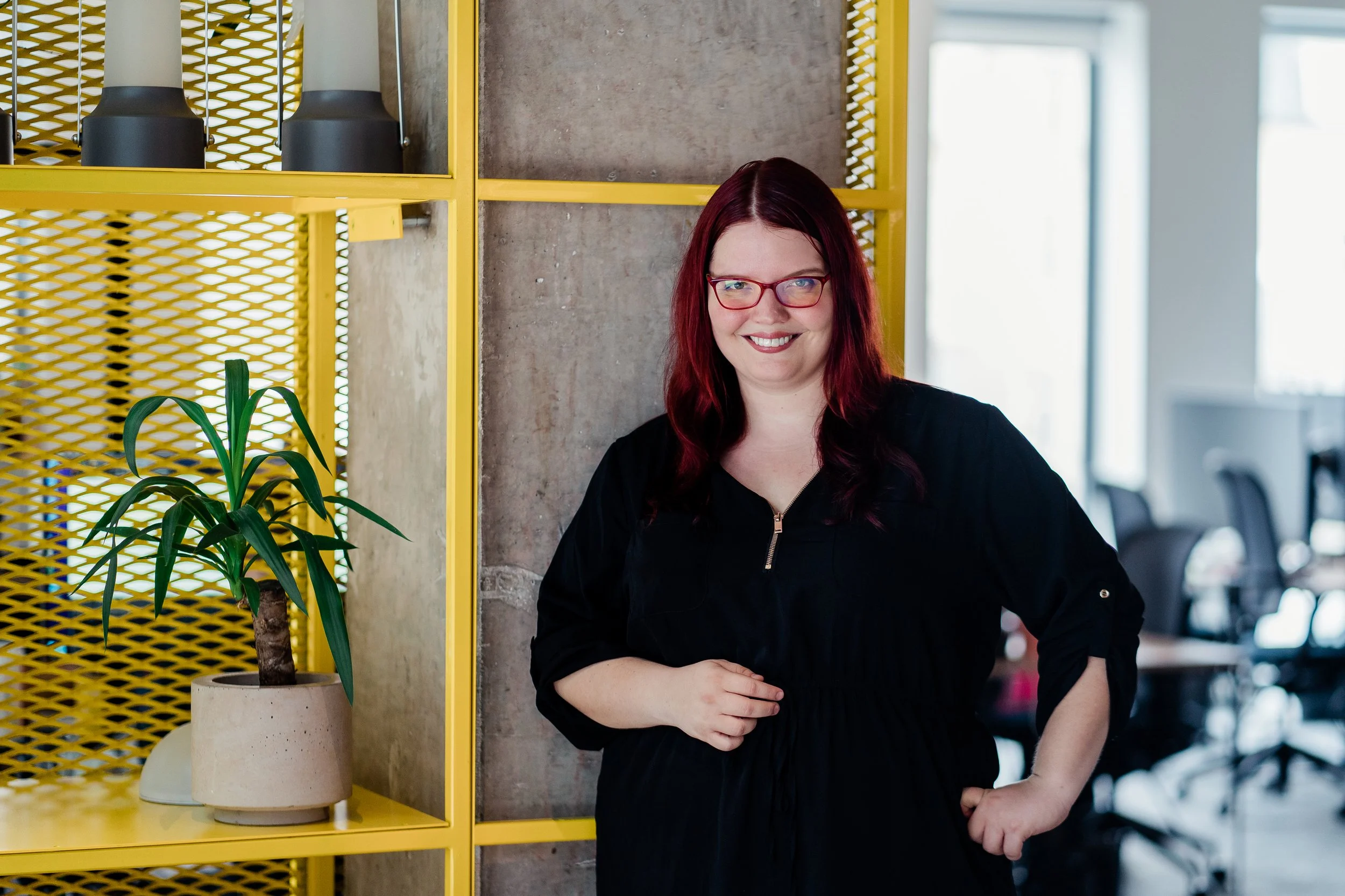 A woman with red hair, glasses, wearing a black dress, smiling and standing next to a yellow metal shelf with indoor plants in office. Professional business headshot photo in Zürich, Zug, Bern, Lucerne, Basel, St. Gallen and across Switzerland.