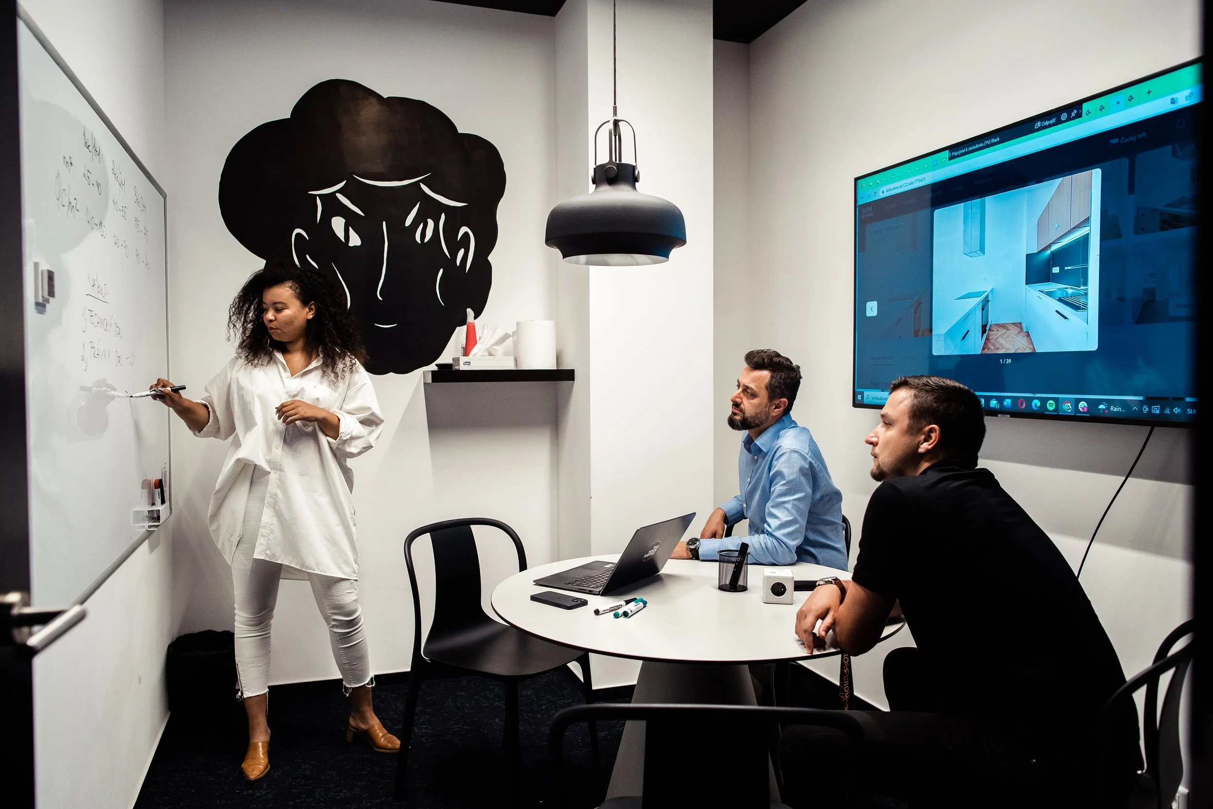 Woman in white shirt writing on a whiteboard while two men sit at a table watching her, in a modern office room with a large wall art of a woman's face and a computer screen displaying a kitchen design. Office life photoshoot Switzerland.