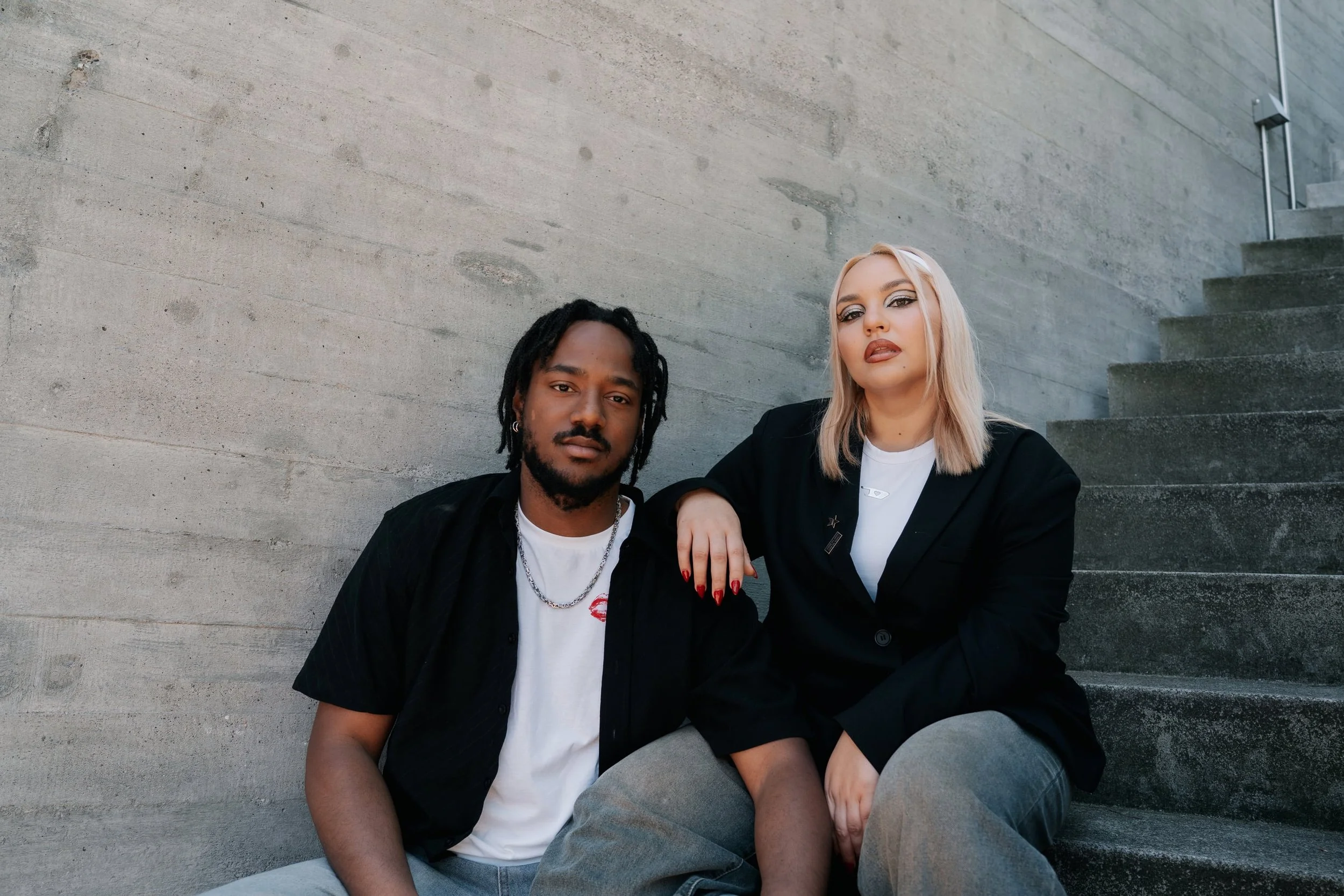 A fashionable couple sitting on concrete stairs against a concrete wall, posing confidently for the camera.