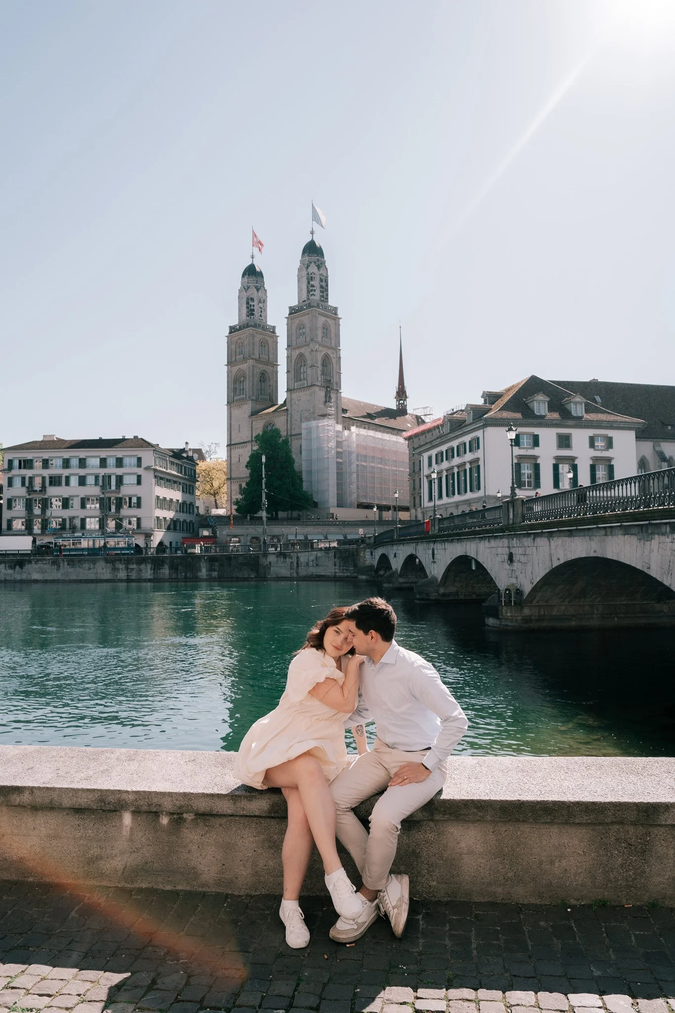 A couple sitting on a stone bench by a river, with a historic church and buildings of Zurich city centre in the background.