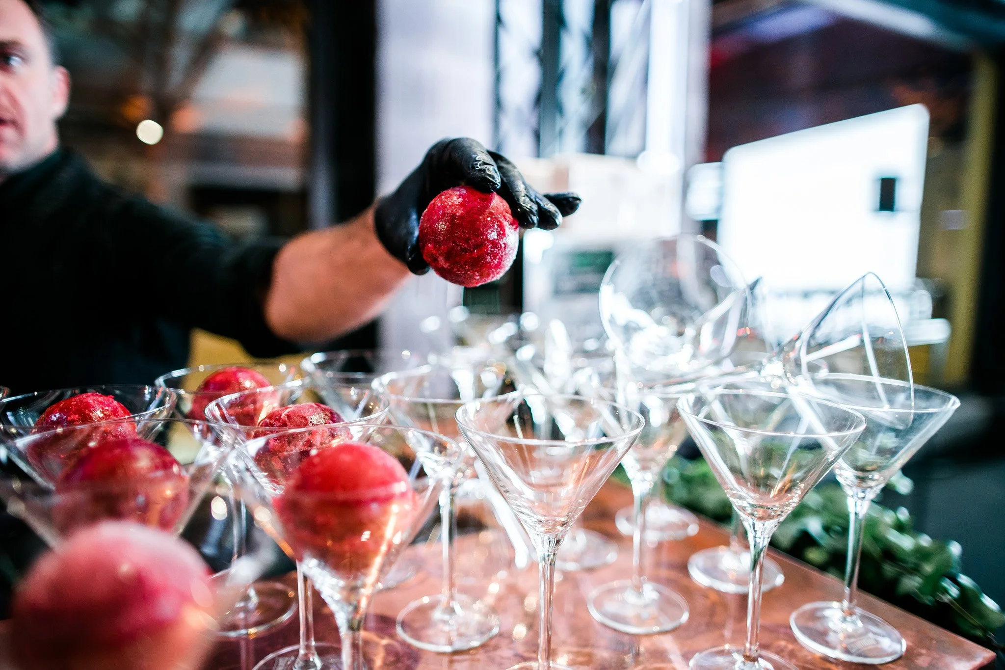 A person wearing black gloves holding a red frozen drink or dessert over a table filled with empty and filled martini glasses. Catering and food photography in Zürich, Zug, Bern, Lucerne, Basel, St. Gallen and across Switzerland.