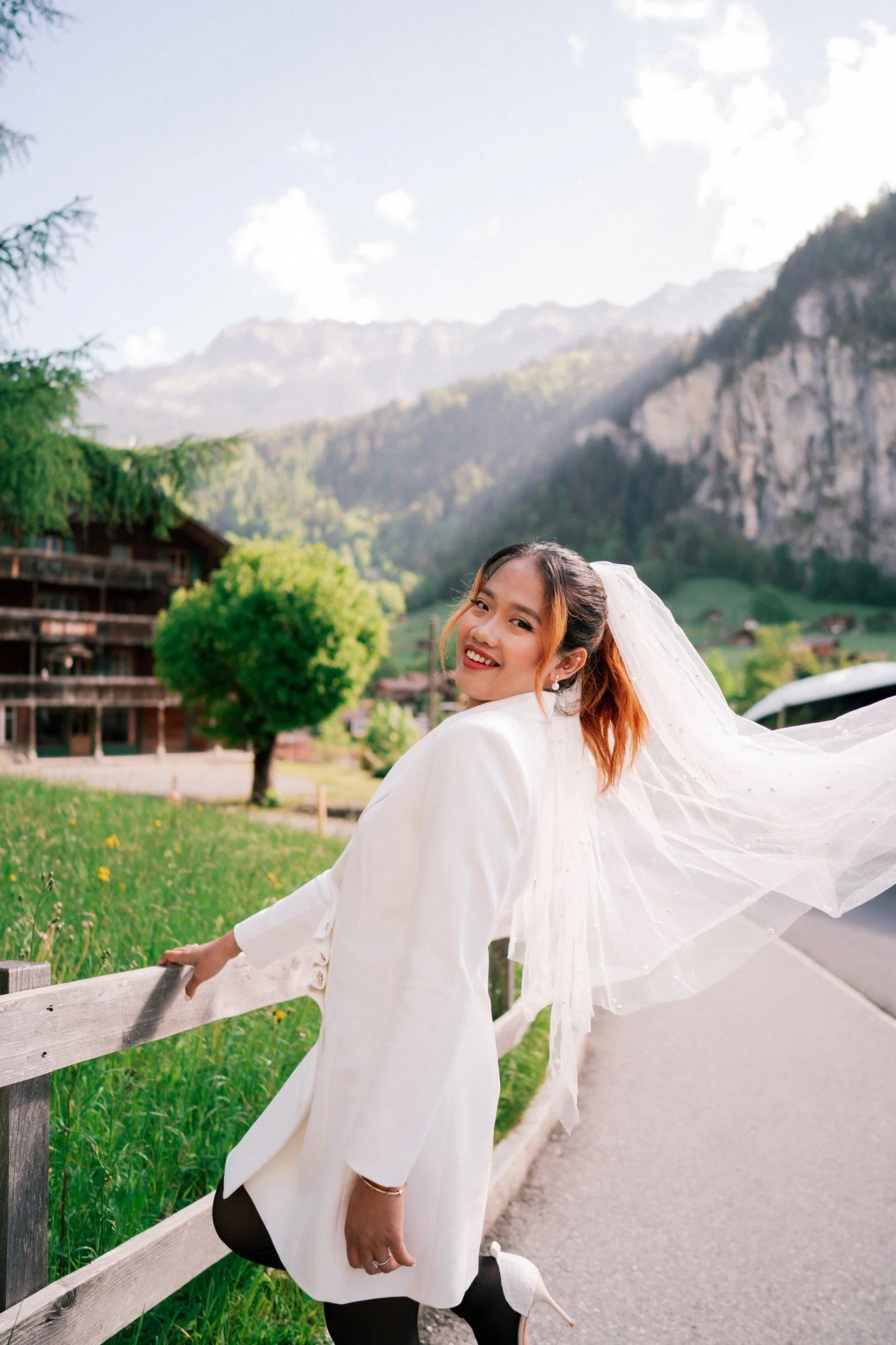 Engagement photoshoot with white dress and veil smiling outdoors on a road with mountains of Lauterbrunnen Switzerland and a wooden building in the background.