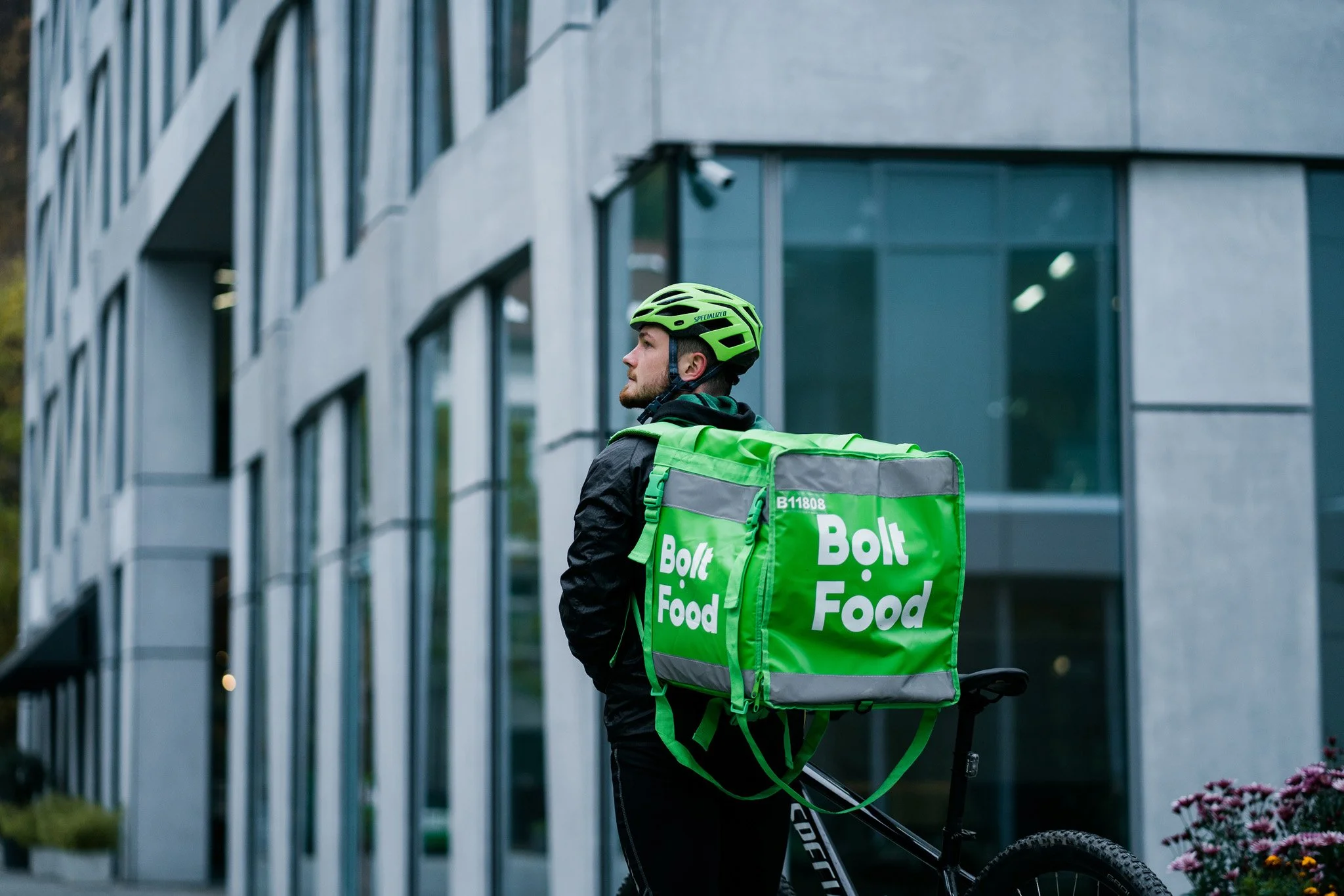 A man wearing a green helmet and black jacket standing next to a bicycle while carrying a large green insulated food delivery bag with the logo 'Bolt Food' on it, in front of a modern building with large glass windows. Advertising photographer Zurich