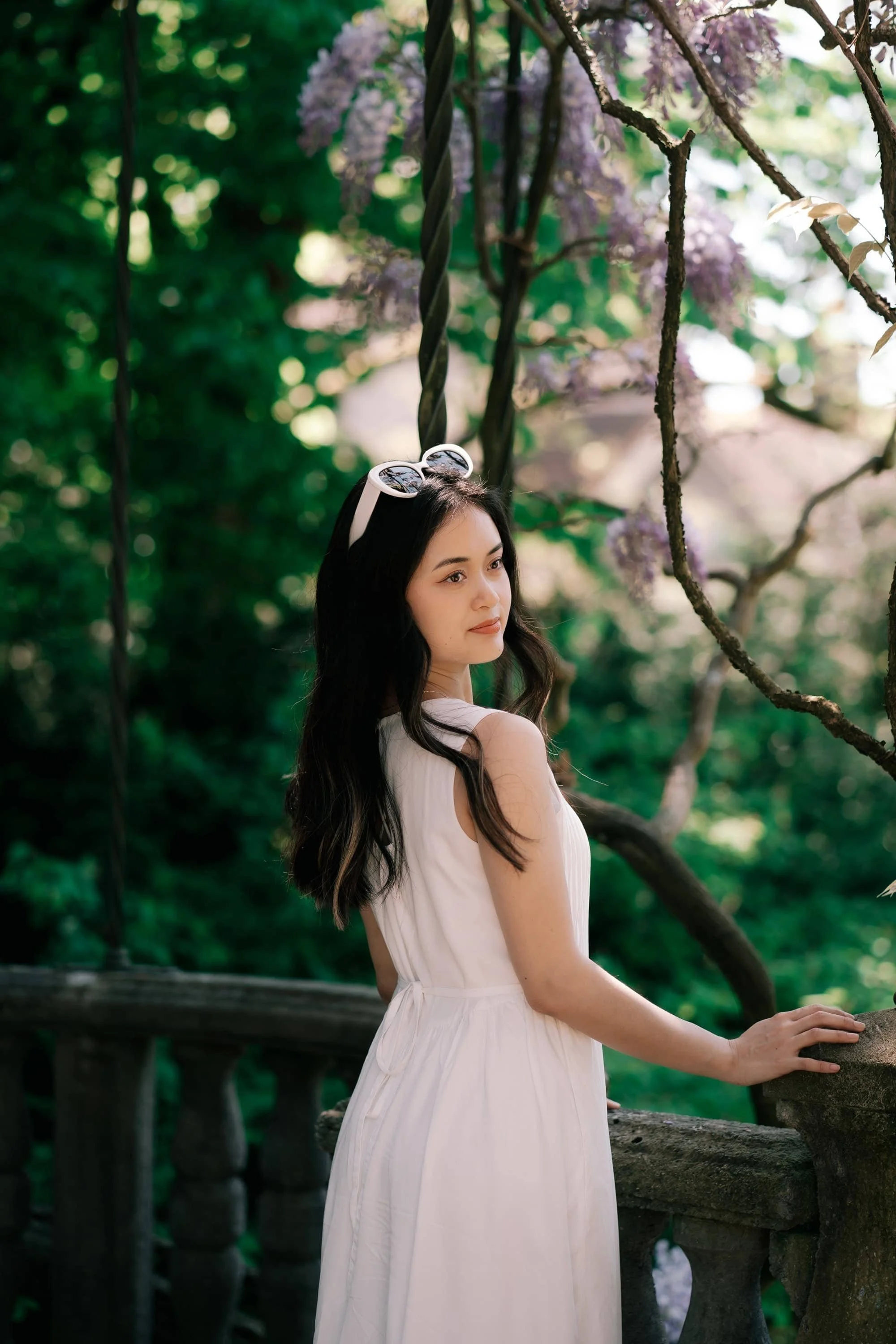 A woman in a white dress standing on a stone bridge in a lush garden with purple flowers and green foliage. Villa Patumbah photoshoot in Zurich.