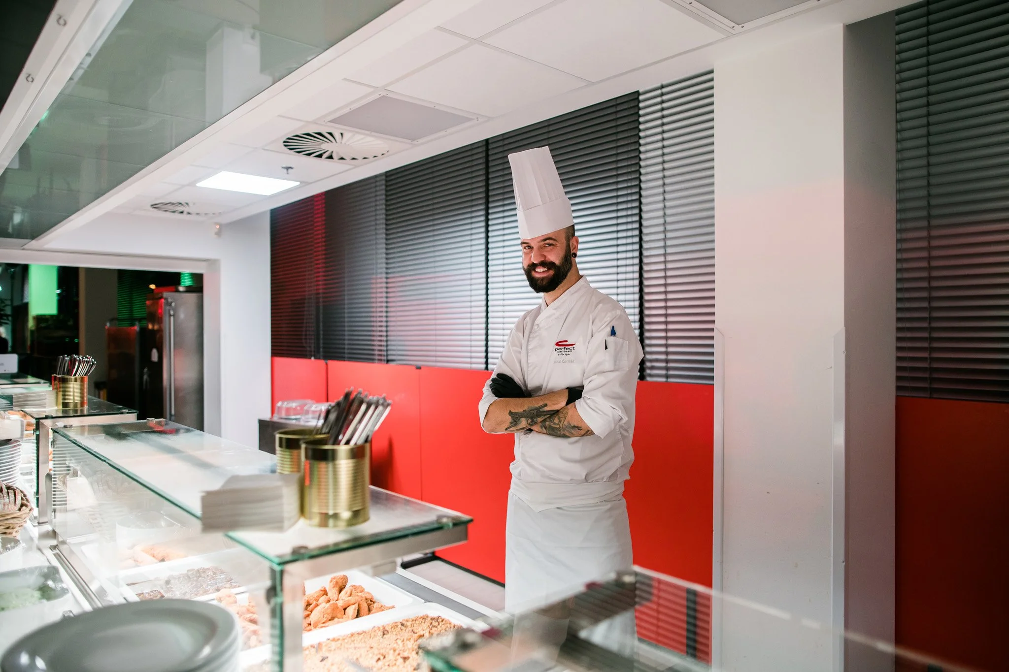 A smiling chef with a beard and tattoos, wearing a tall chef's hat and white uniform, standing with arms crossed behind a glass food display counter in a modern kitchen or restaurant setting. Catering, culinary and food photography in Switzerland.
