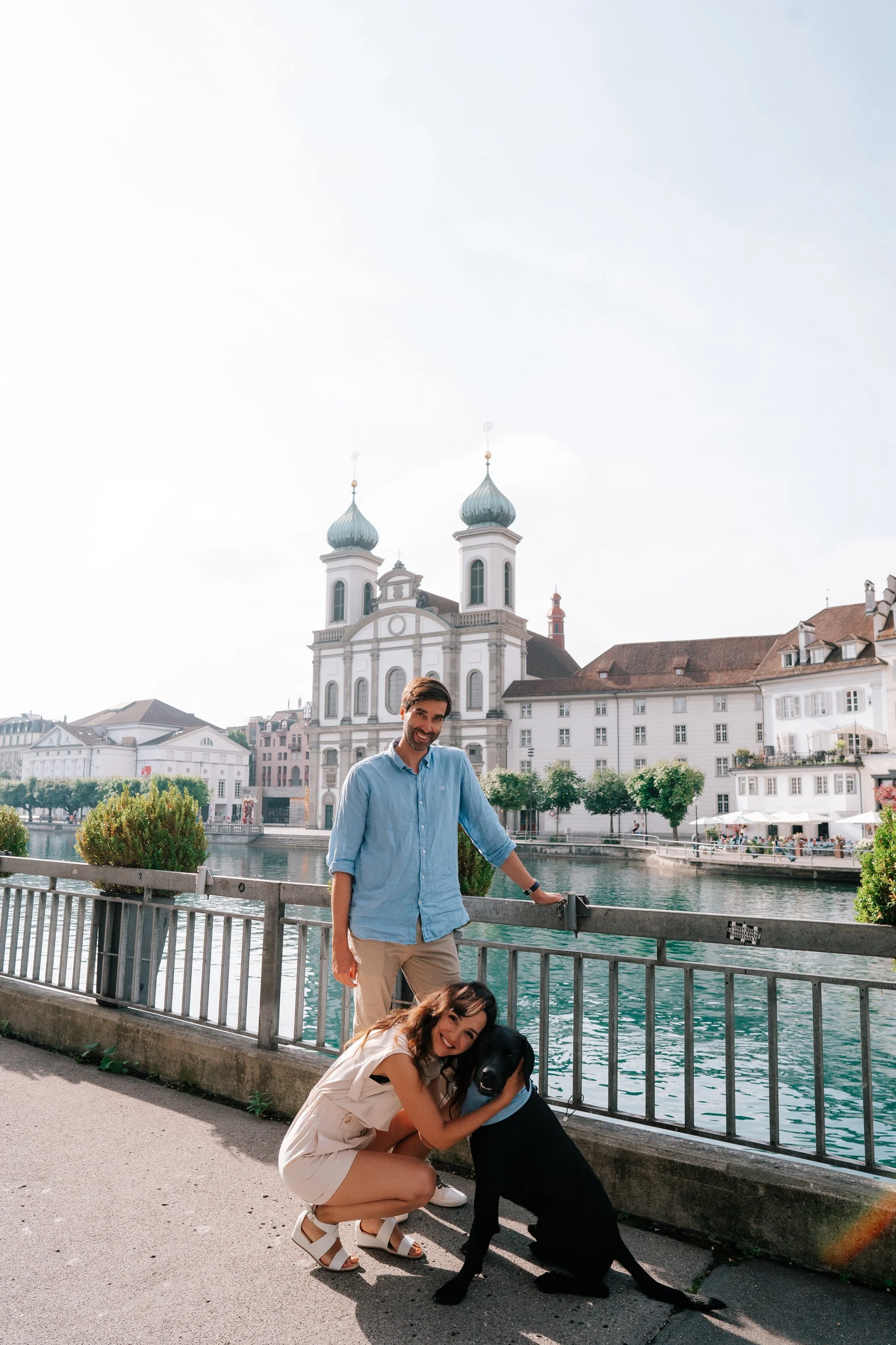A man and a woman with a black dog standing by a river in Lucerne Switzerland, with historical European buildings in the background.