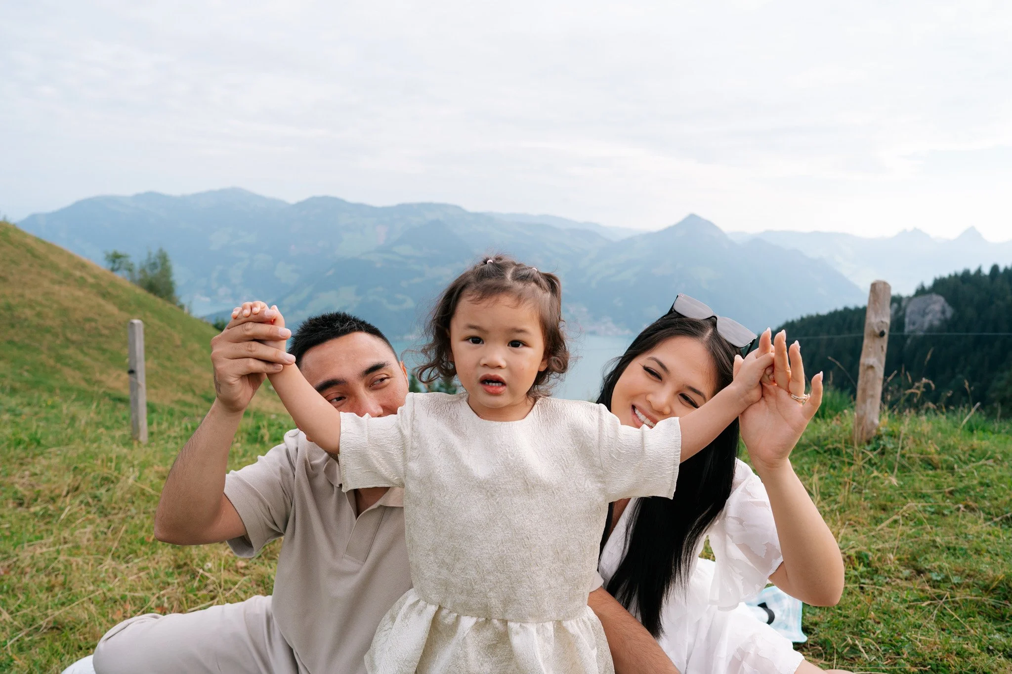 A family of three, including a man, a woman, and a young girl, outdoors on a grassy hillside with mountains in the background. The girl is centered with her arms raised, and the parents are smiling nearby. Family photoshoot in Switzerland.
