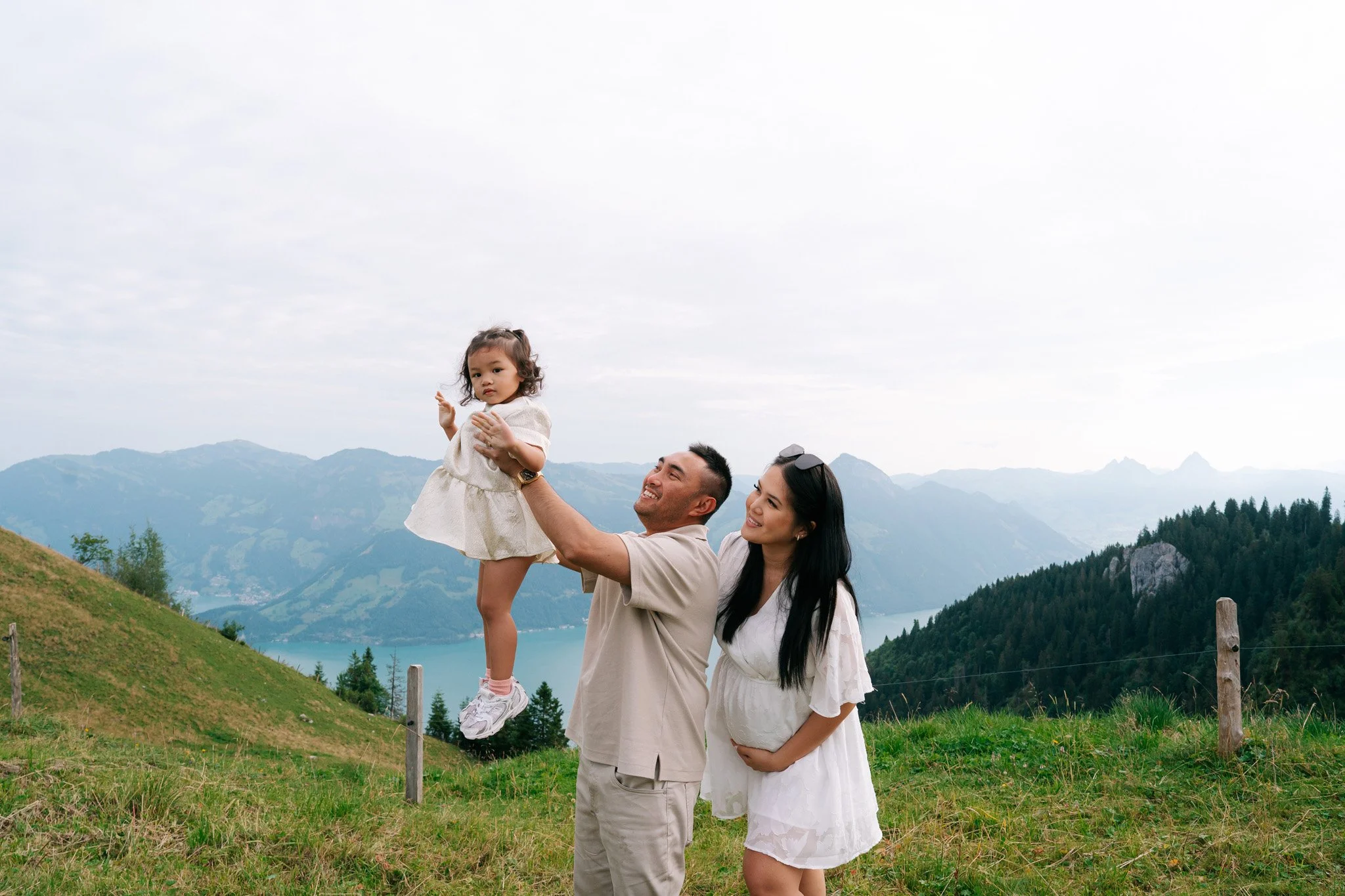 Family enjoying time outdoors in Swiss Alps on a grassy hillside with mountains and lake in the background, the father lifting a young girl while the mother stands nearby and smiles. Family photoshoot in Switzerland.