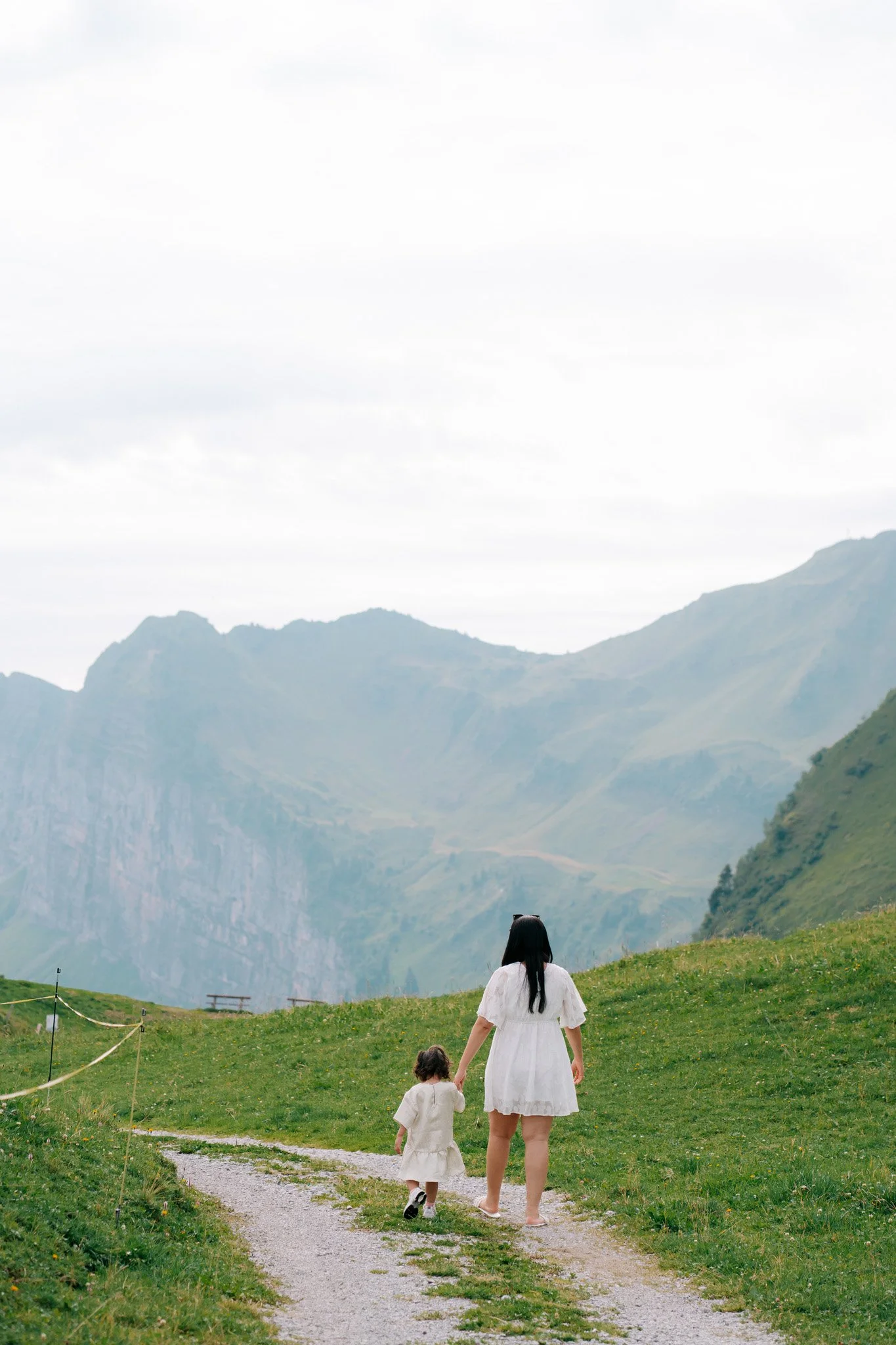 A woman and a young girl walking hand in hand on a grassy trail in a mountainous area with cloudy sky in the background. Family photoshoot in Switzerland.