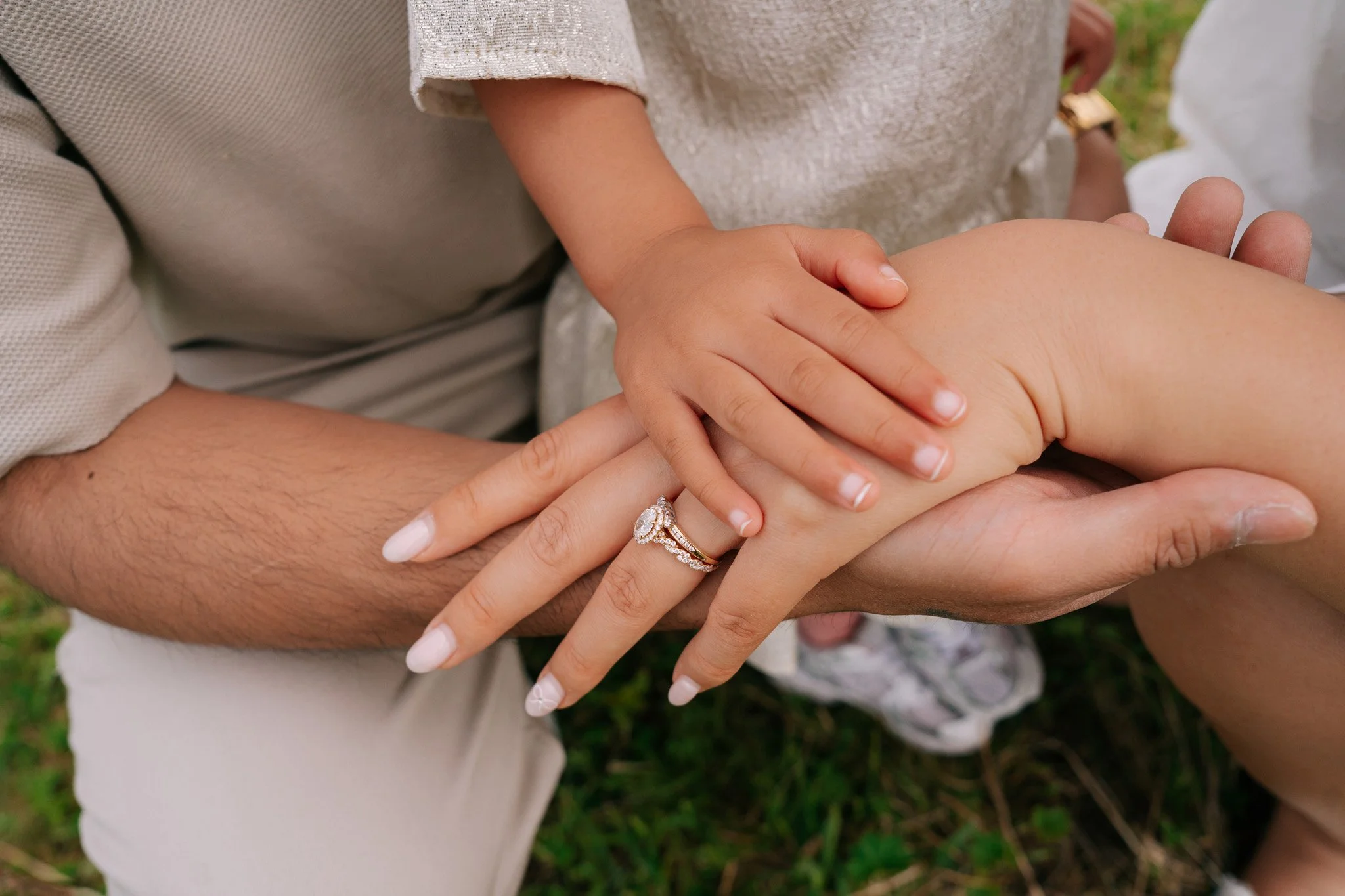 A person wearing a ring holds a child's hand, both resting on a person's knee in an outdoor setting with grass. Family photoshoot in Switzerland.