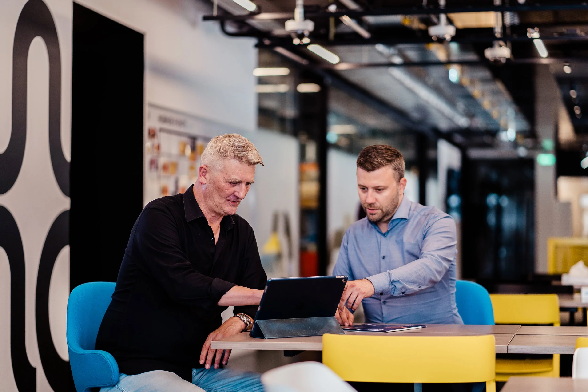 Two men sitting at a table in an office environment, looking at a tablet device together having a meeting. Modern employer branding photography in Zürich, Zug, Bern, Lucerne, Basel, St. Gallen and across Switzerland.