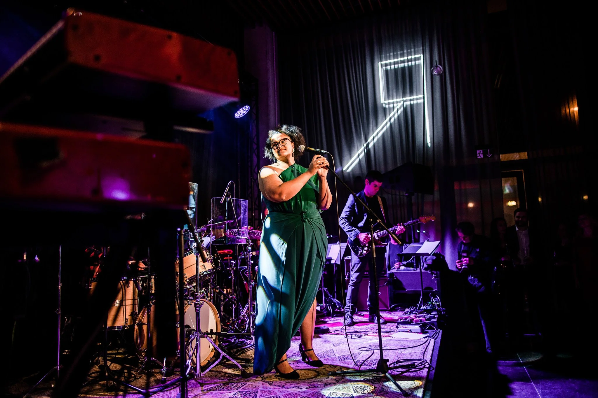 A woman in a green dress singing into a microphone on stage with a band, with neon light artwork in the background. Concert photography in Zürich, Zug, Bern, Lucerne, Basel, St. Gallen and across Switzerland.