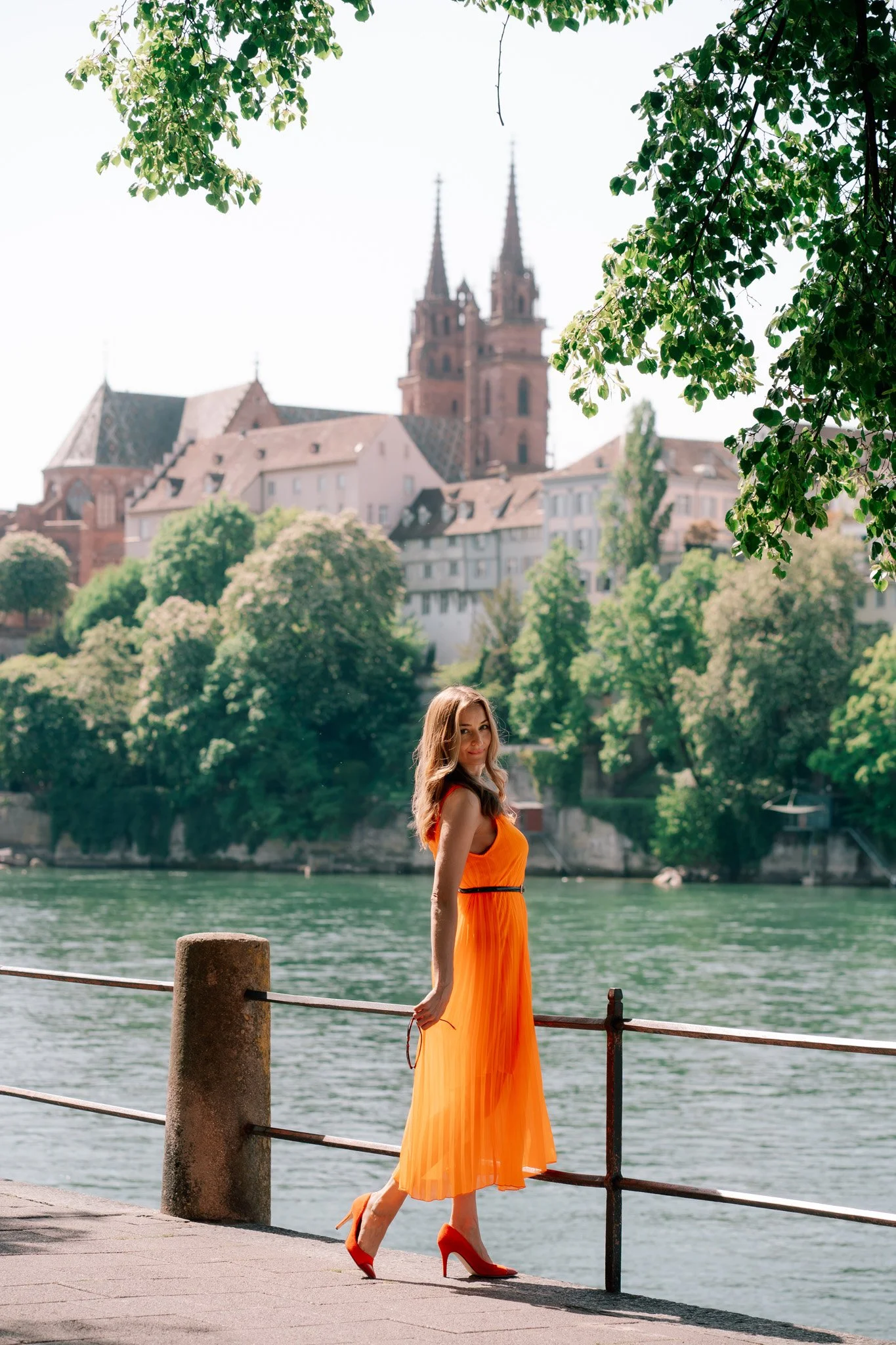 Solo portrait photoshoot woman in an orange dress and red high heels walking along a riverside promenade in Basel, Switzerland and church in the background.