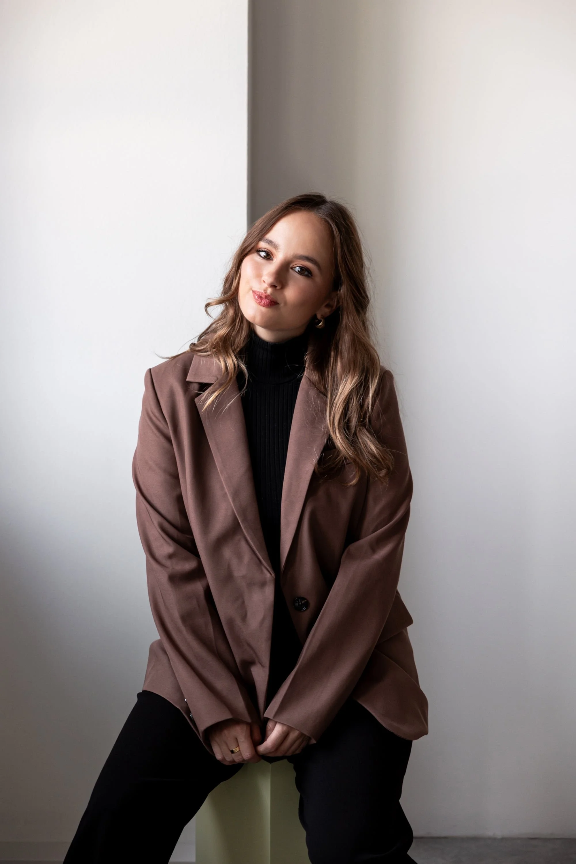 Switzerland photographer with wavy brown hair wearing a brown blazer and black turtleneck sitting in front of a plain wall.